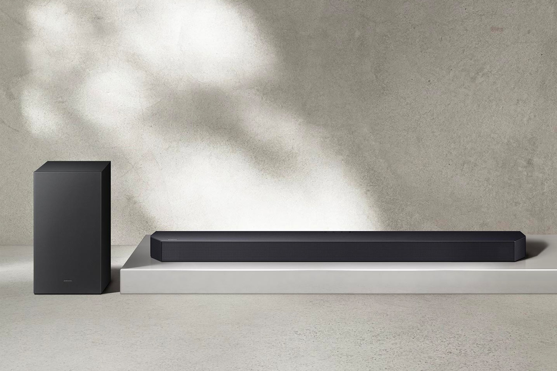 Eine Soundbar und ein Subwoofer stehen auf einer weißen Plattform vor einer strukturierten grauen Wand. Schatten und Licht schaffen eine minimalistische Szene.