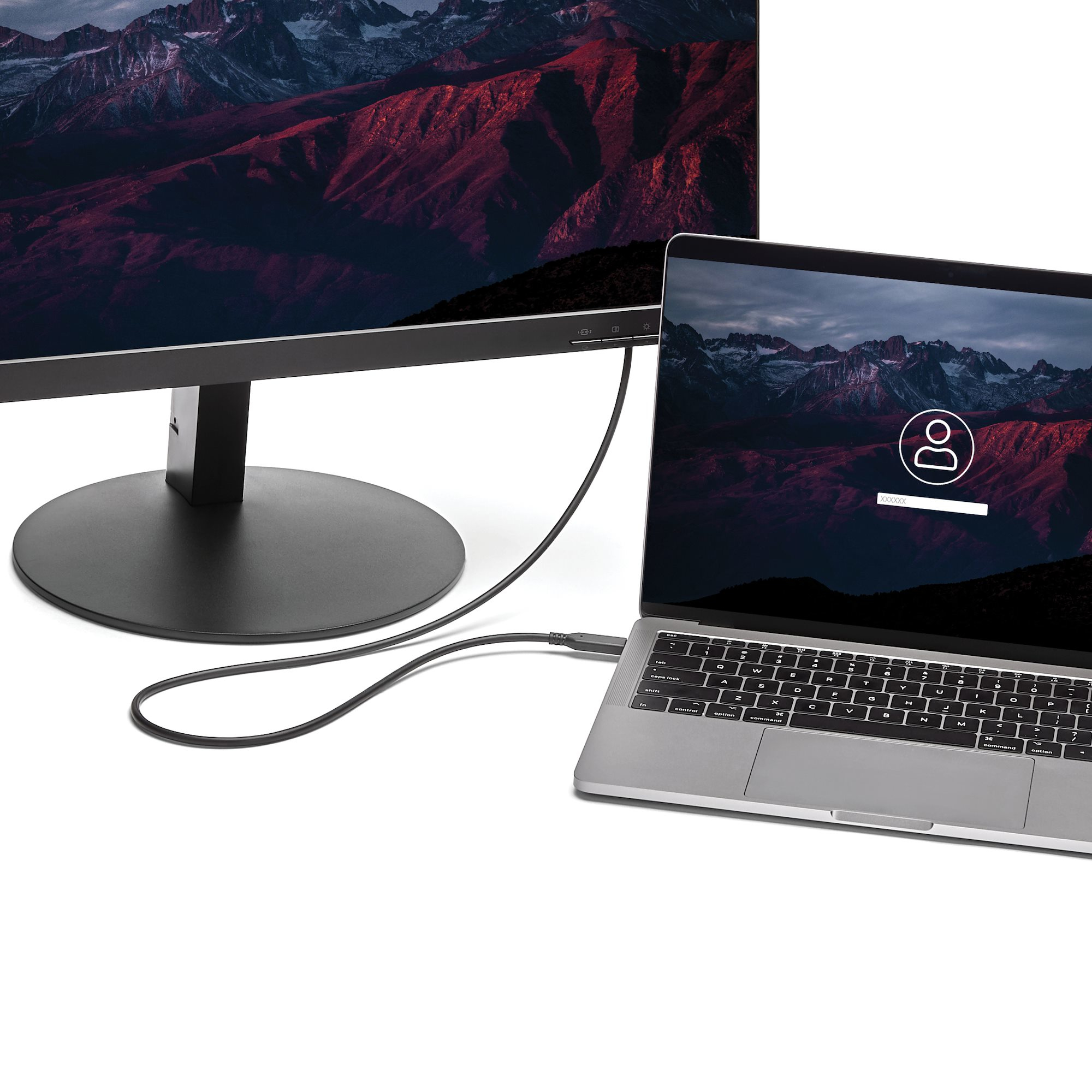 Un portátil conectado a un monitor por cable negro. Ambas pantallas muestran una escena de montaña. El portátil tiene una pantalla de inicio de sesión.