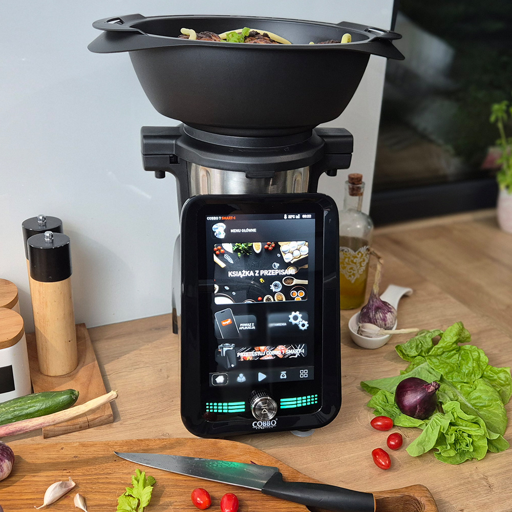 Scena kuchenna z robotem kuchennym, warzywami i nożem na drewnianej desce. Ekran pokazuje opcje menu.