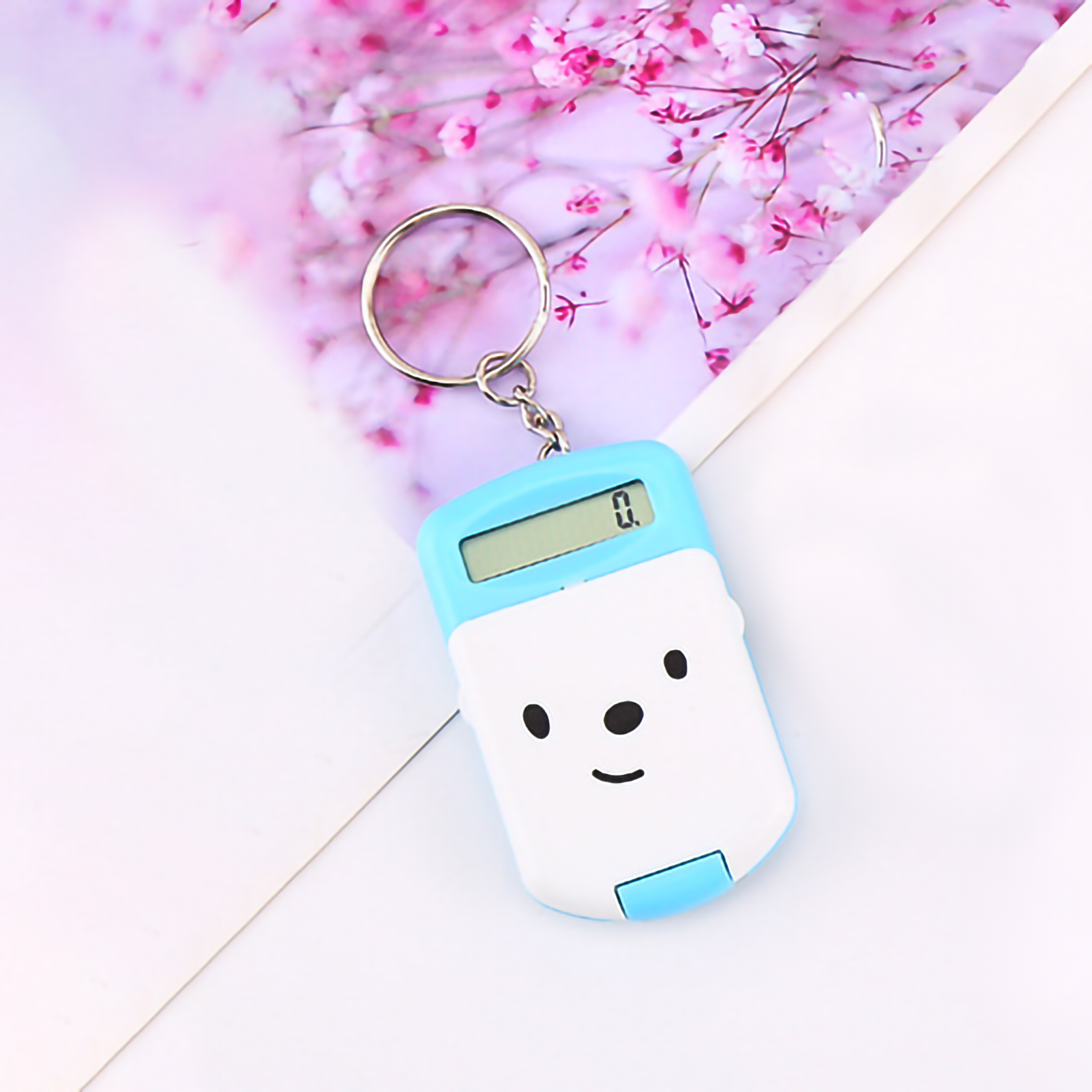 Ein blau-weißer Taschenrechner-Schlüsselanhänger mit Smiley-Gesicht liegt auf weißem Untergrund mit rosa Blumenhintergrund.