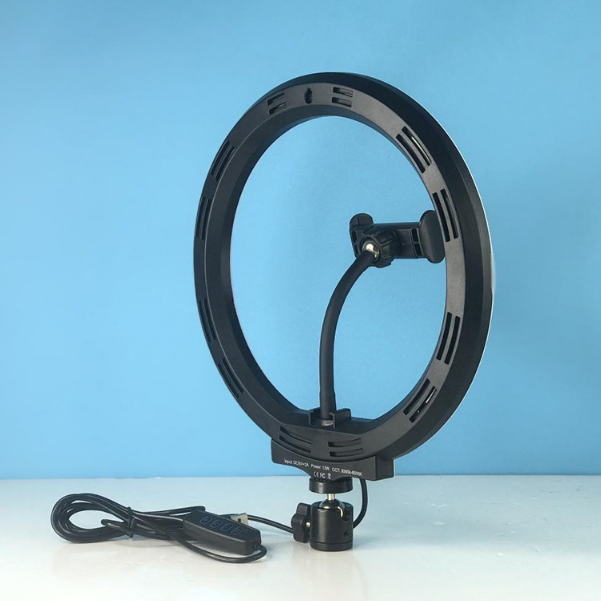 Ein schwarzes Ringlicht mit flexiblem Arm und Telefonhalter steht auf weißem Untergrund vor blauem Hintergrund.