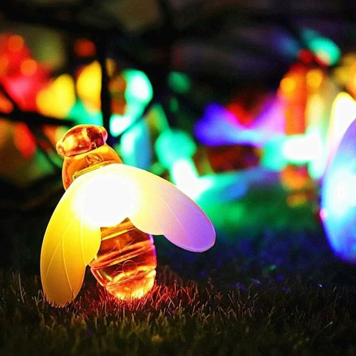 Ein leuchtendes bienenförmiges Licht steht auf Gras, umgeben von bunten, verschwommenen Lichtern im Hintergrund, was eine festliche Szene schafft.