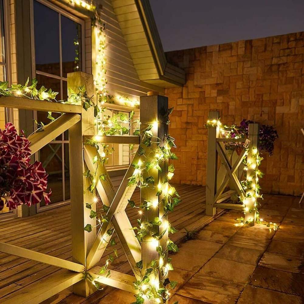 Holzdeck mit Lichterketten und Efeu. Ein Gebäude mit Holzzaun. Blumen im Vordergrund. Nachtszene.
