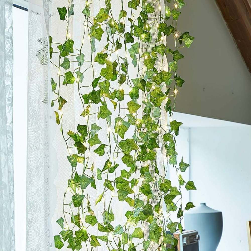 Grüner Efeugirlande mit Feenlichtern, die vor einem weißen Vorhang hängen. Im Hintergrund ist eine blaue Vase zu sehen.