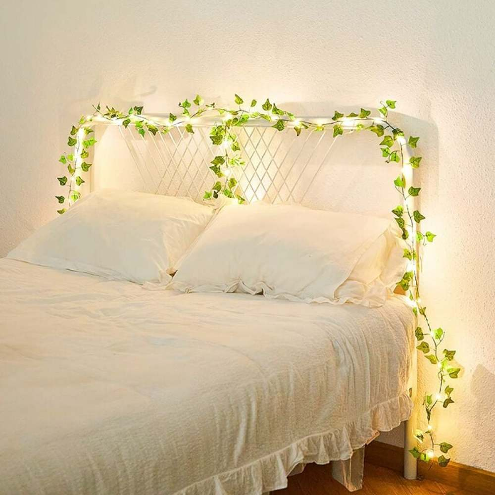 Bett mit weißer Bettwäsche, Kopfteil mit grünen Blätterschnur-Lichtern. Die Wand ist hell. Sanfte Beleuchtung.