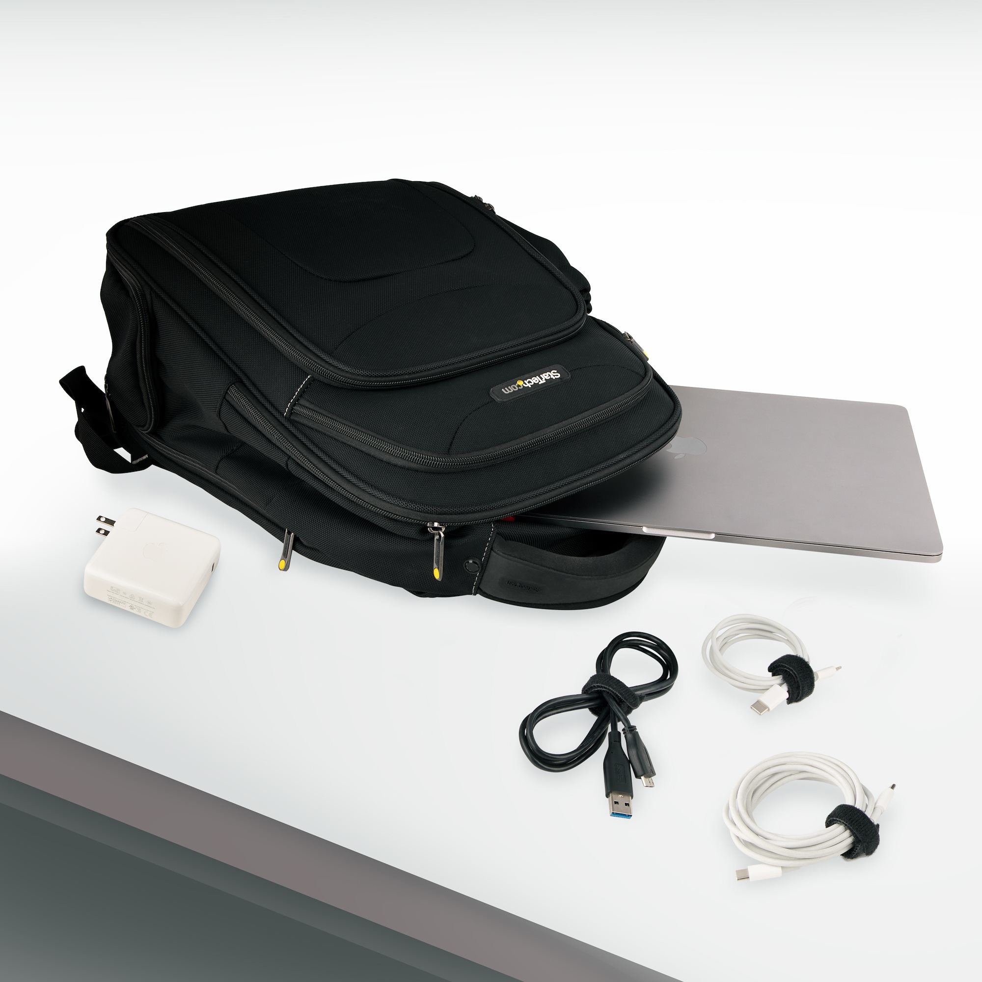 Zaino nero con laptop, caricatore bianco e due cavi USB su una superficie bianca.