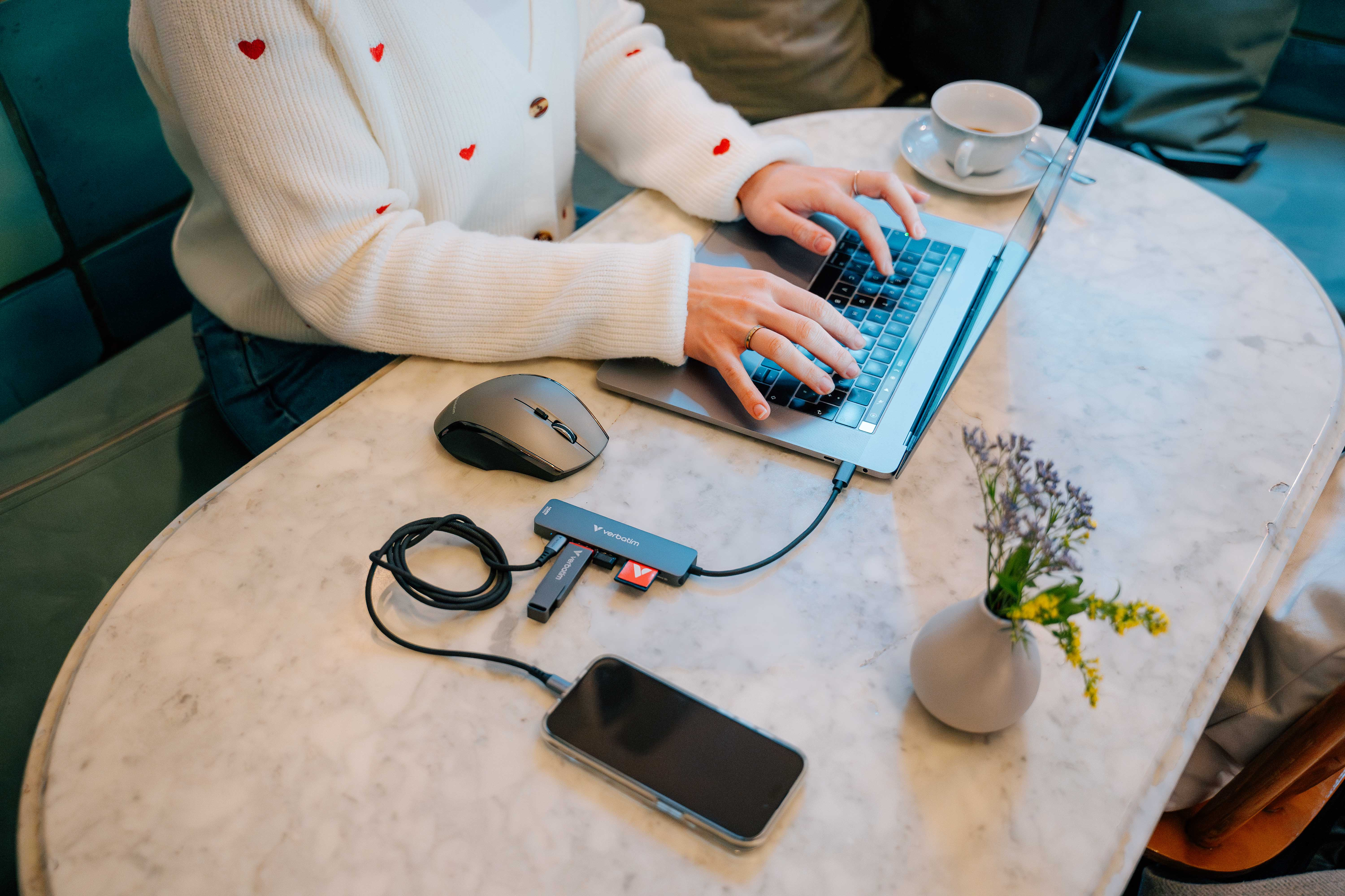Eine Person tippt auf einem Laptop an einem Tisch. Maus, Telefon und Vase mit Blumen sind ebenfalls vorhanden.