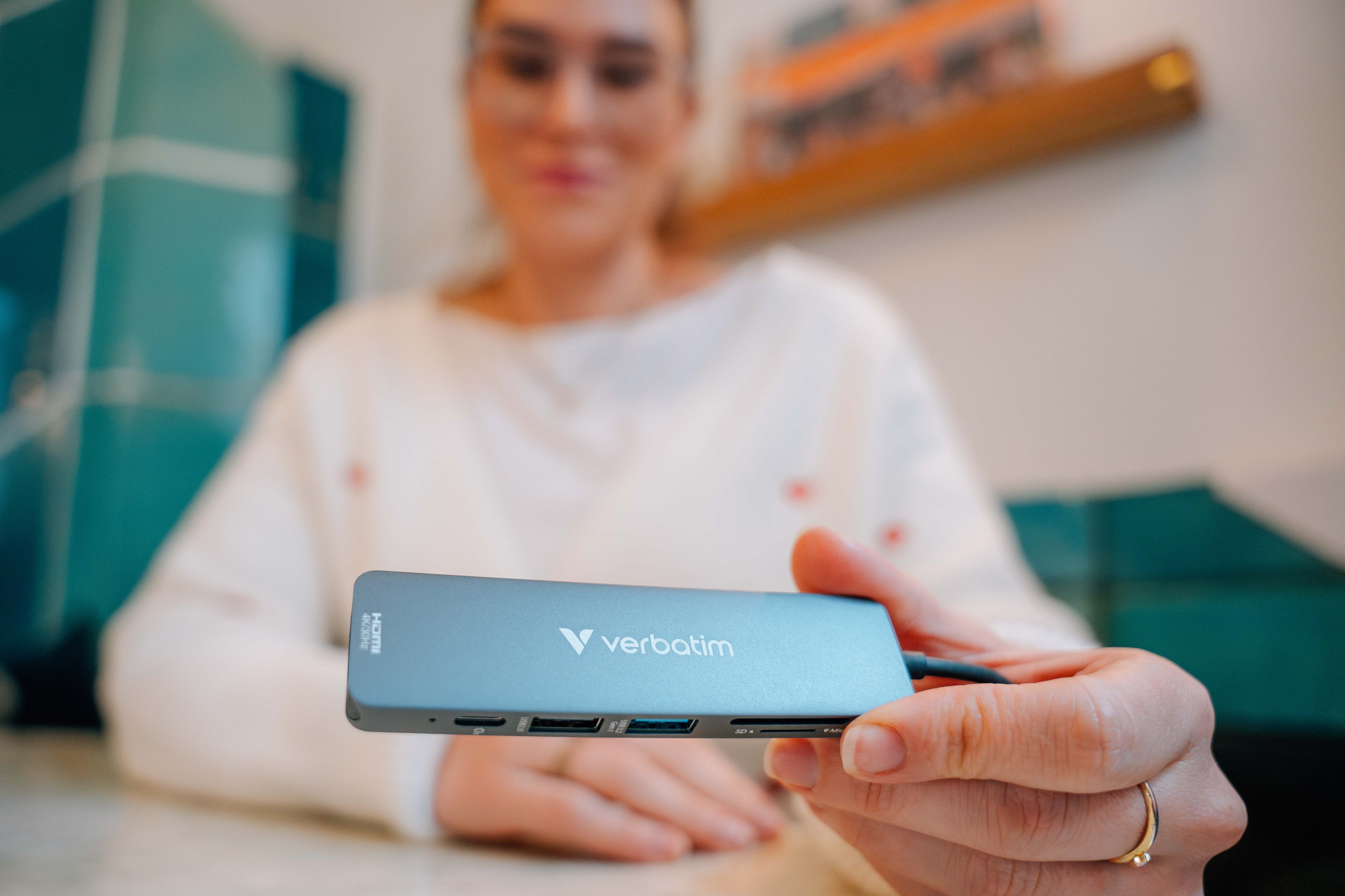 Eine Person hält einen Verbatim USB-Hub. Der Hub ist silberfarben und hat verschiedene Anschlüsse. Die Person ist unscharf.