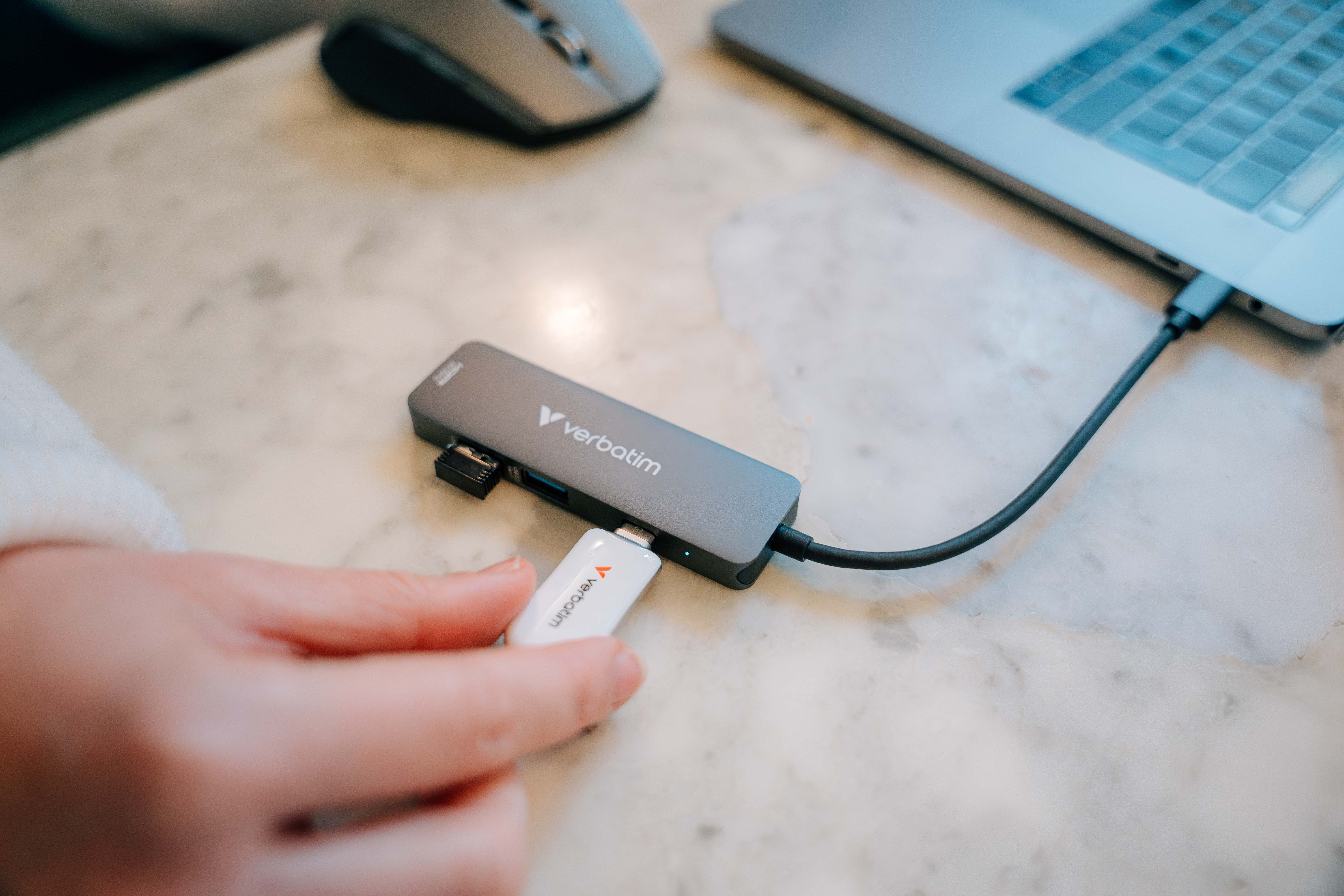 Hand steckt Verbatim USB-Laufwerk in einen grauen Hub, der an einen Laptop auf einer Marmoroberfläche angeschlossen ist.