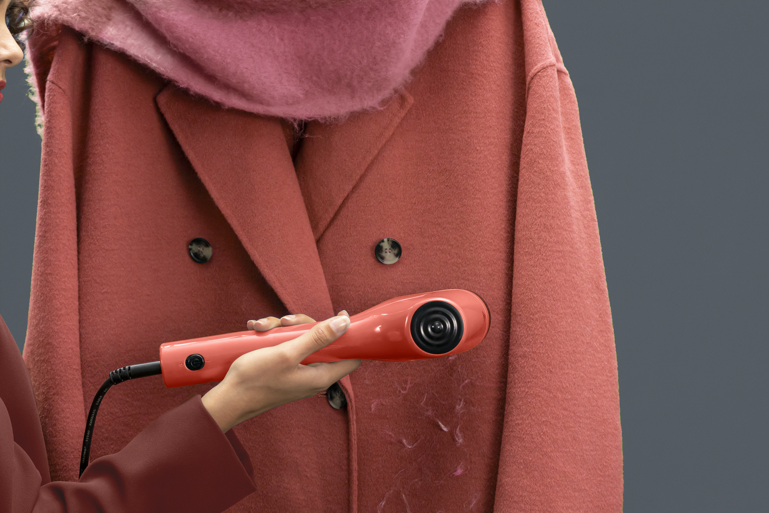 Osoba używa golarki do tkanin na czerwonym płaszczu. Płaszcz wisi na szarym tle. Różowy szalik jest na płaszczu.