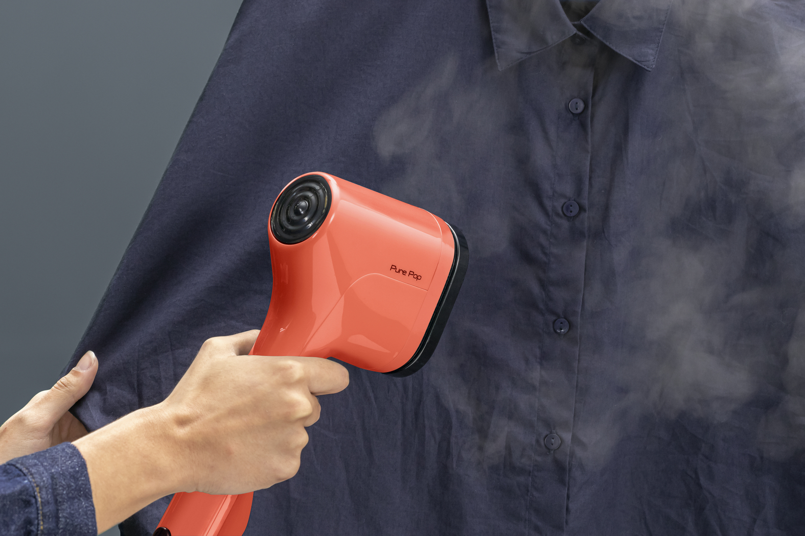 Pomarańczowy parownik do ubrań paruje granatową koszulę. Ręka trzyma parownik, wydobywa się para. Koszula ma guziki.