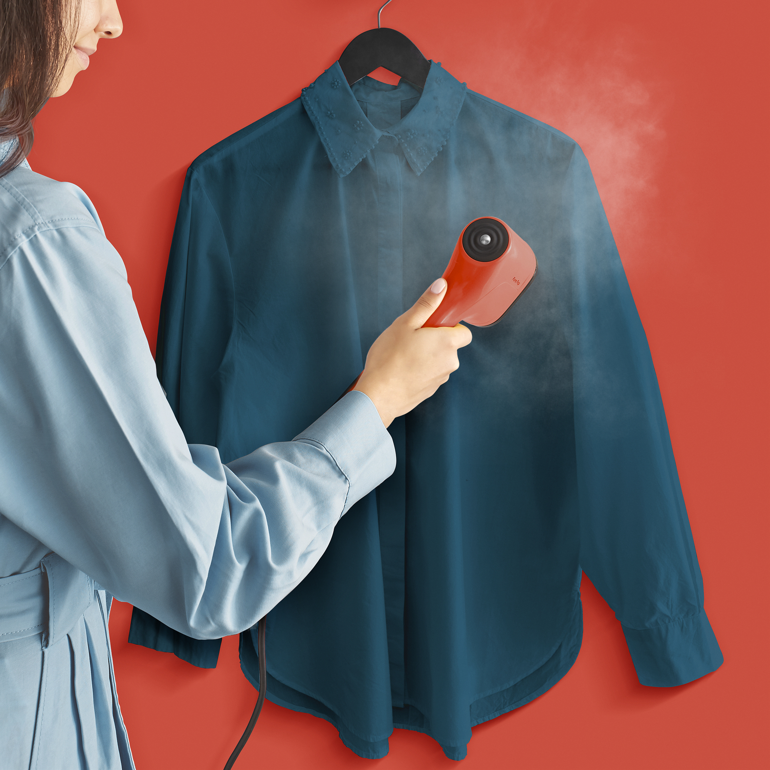 Kobieta prasuje niebieską koszulę pomarańczowym steamerem. Koszula wisi na czarnym wieszaku na czerwonym tle.
