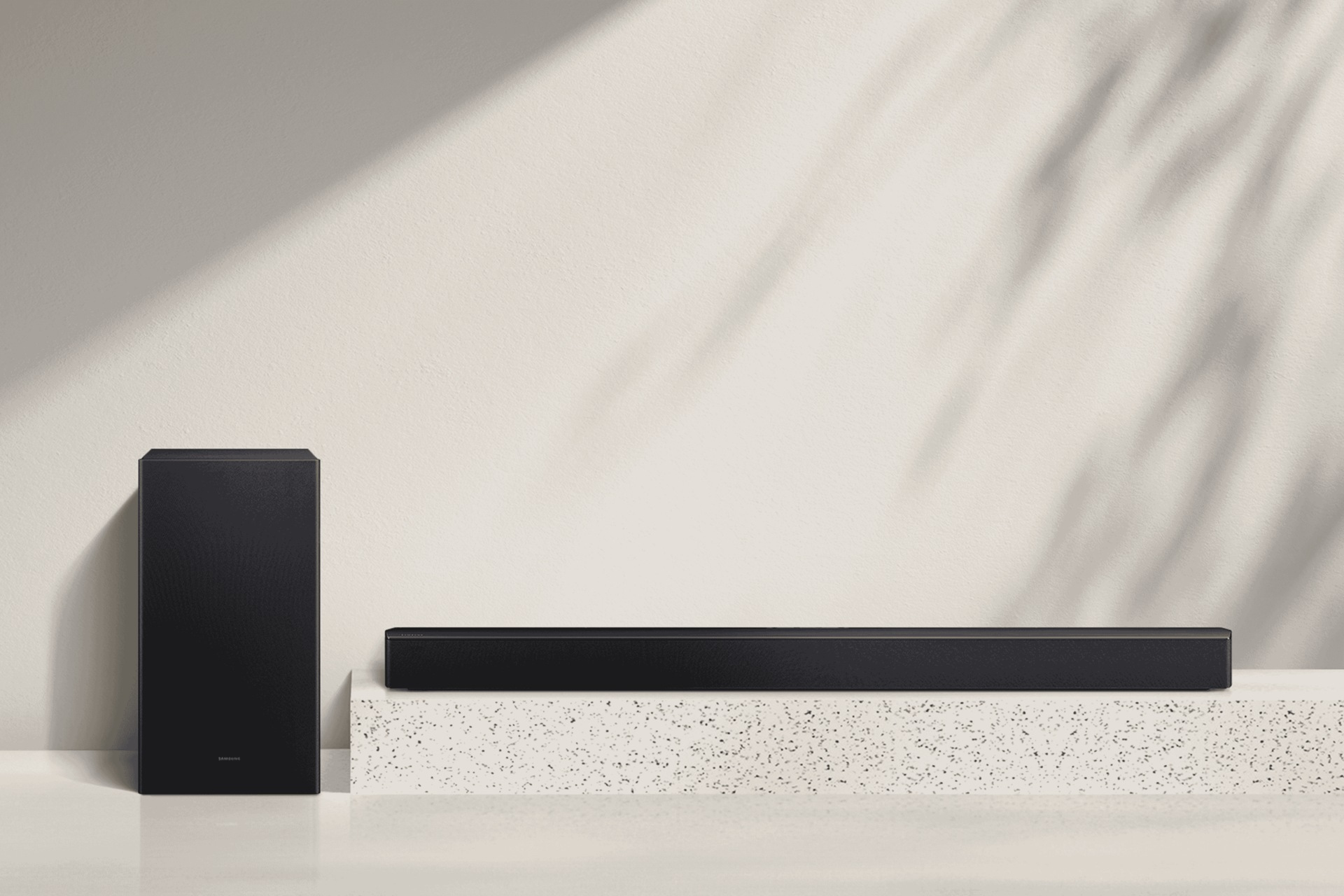 Schwarze Soundbar und Subwoofer auf einer gesprenkelten Plattform vor einer beigen Wand mit Blätterschatten.