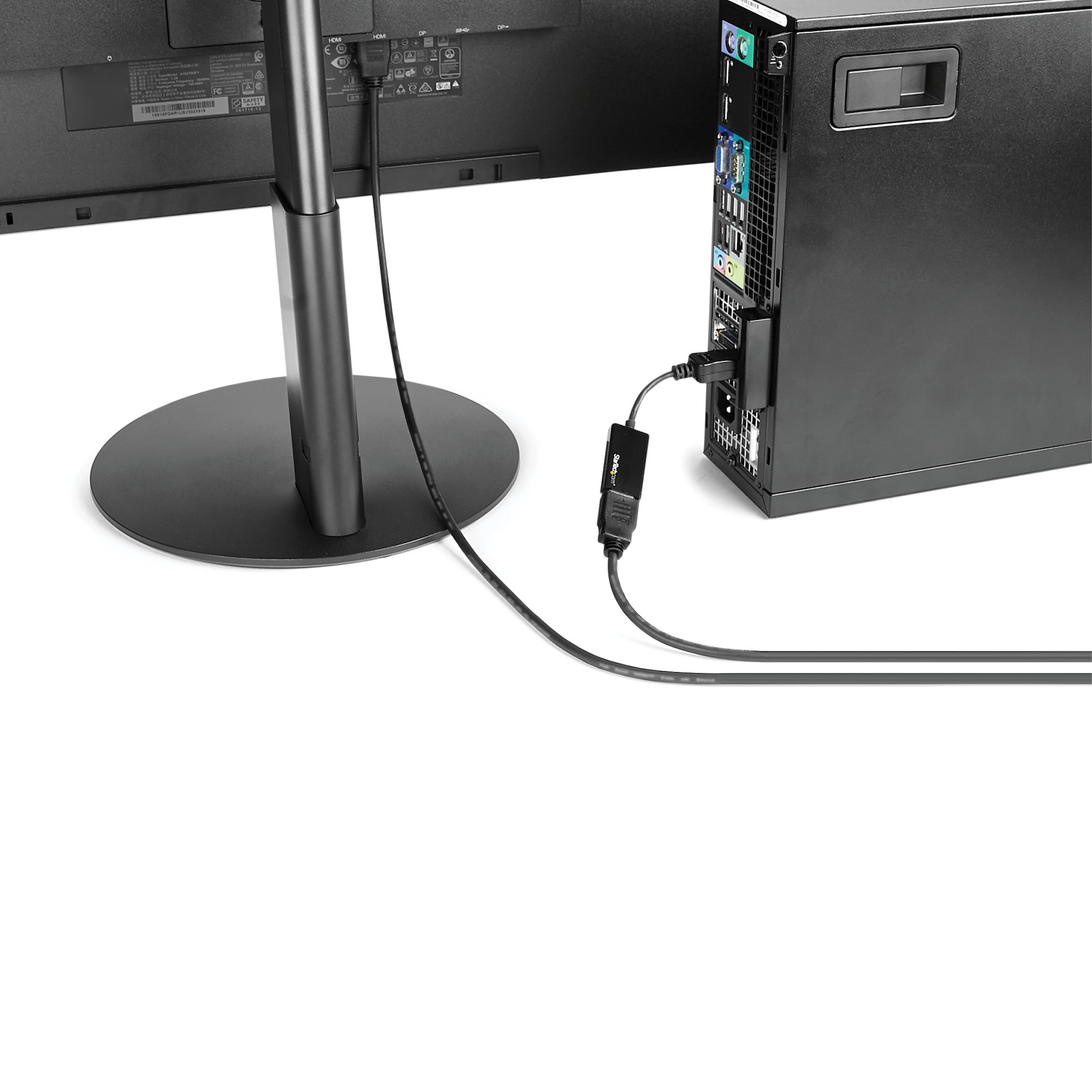 Un monitor de computadora está conectado a una torre de computadora a través de un cable y adaptador negro, sobre un fondo blanco.