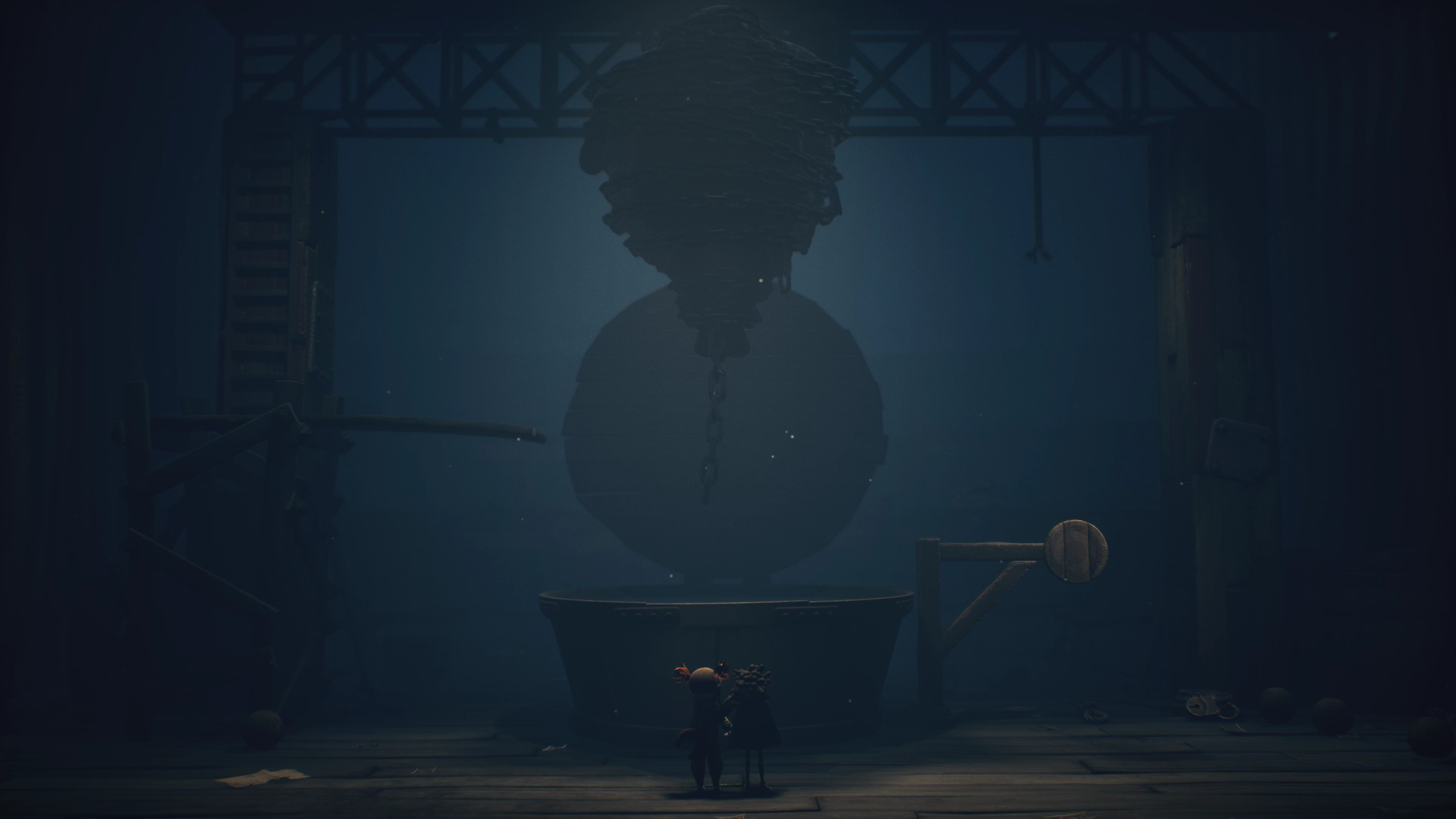 Una scena oscura con un grande oggetto sospeso e due piccole figure. Lo sfondo presenta travi di supporto e un grande contenitore.