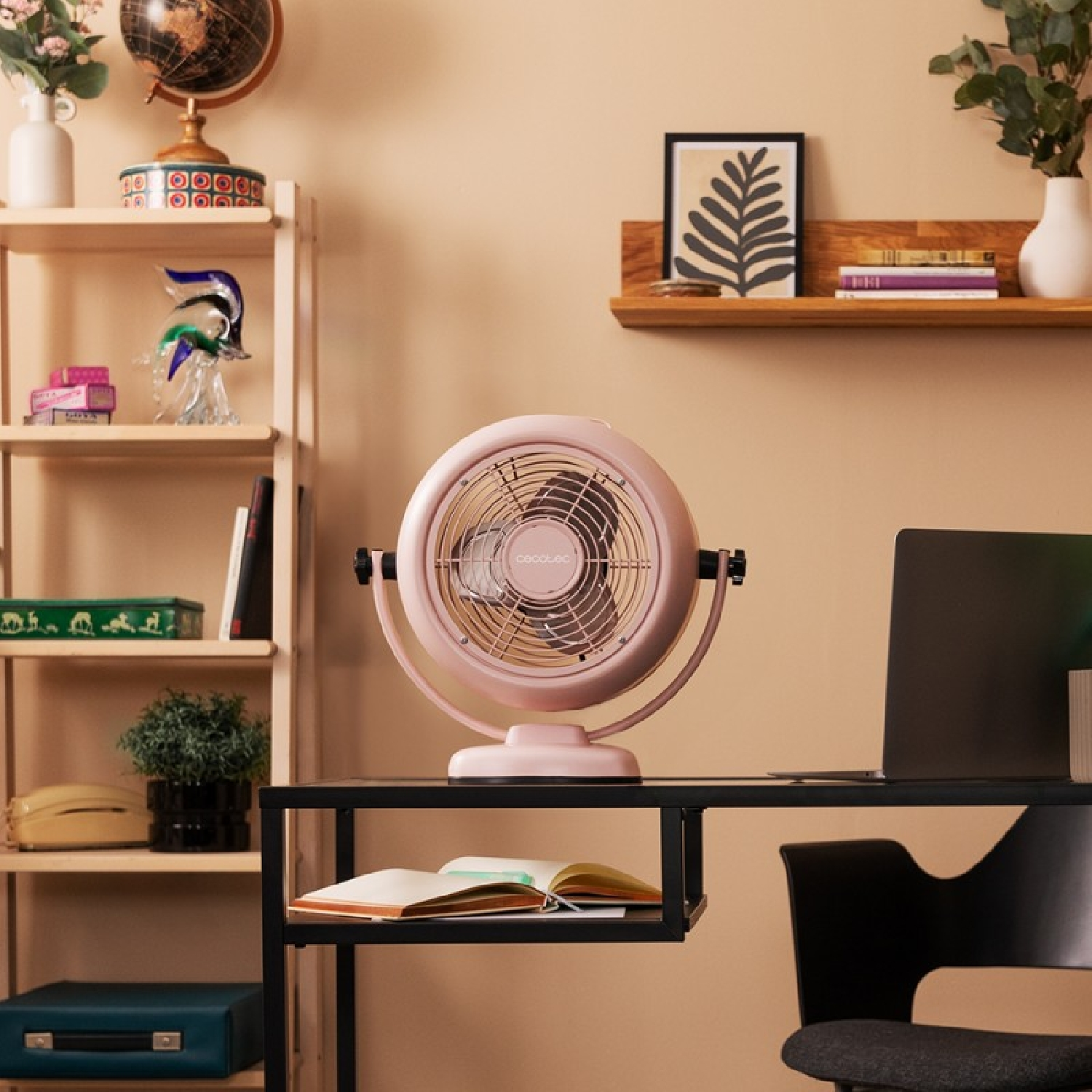 Un ventilatore rosa è su una scrivania nera con un libro aperto. Un laptop, una sedia nera e mensole con decorazioni sullo sfondo.