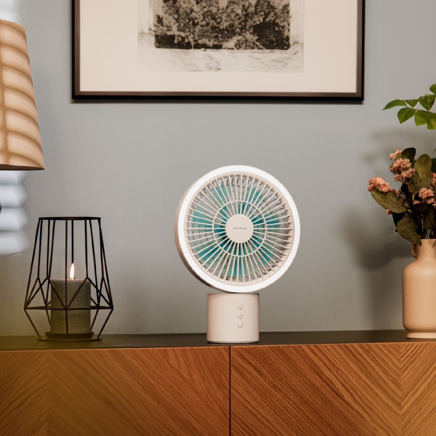 Un ventilador blanco con aspas azules está sobre un mueble de madera. También hay una vela en una jaula negra y un jarrón con flores.