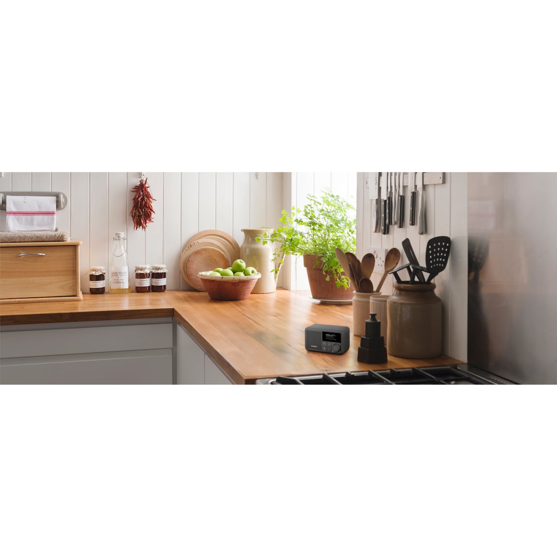 A kitchen counter with a radio, utensils, and jars. Jars of jam, a bread bin, and a bowl of green apples are visible.