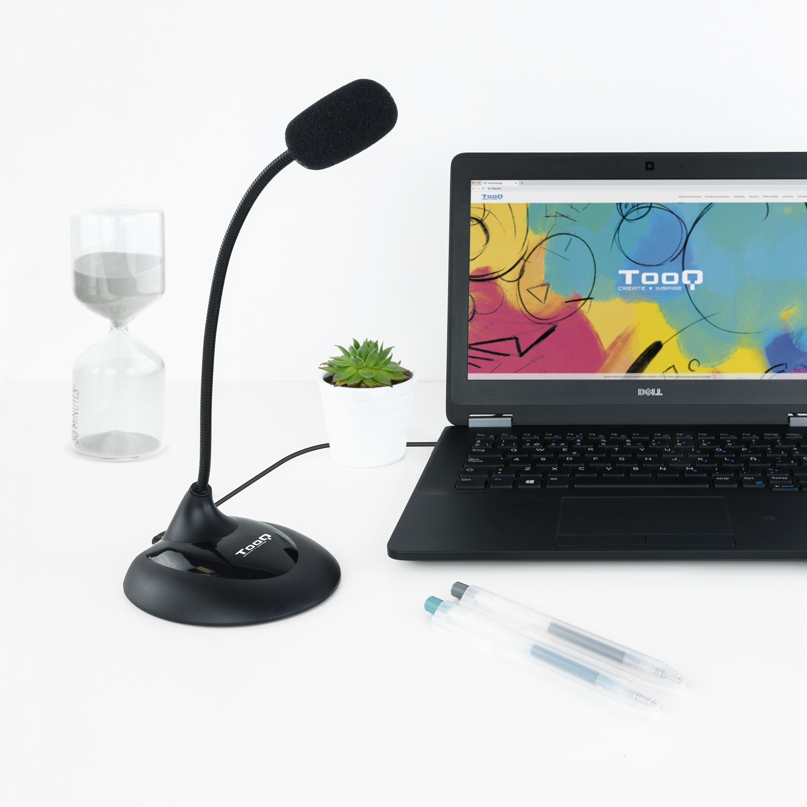 Konfiguracja biurka z mikrofonem, laptopem, sukulentem, klepsydrą i długopisami na białej powierzchni.