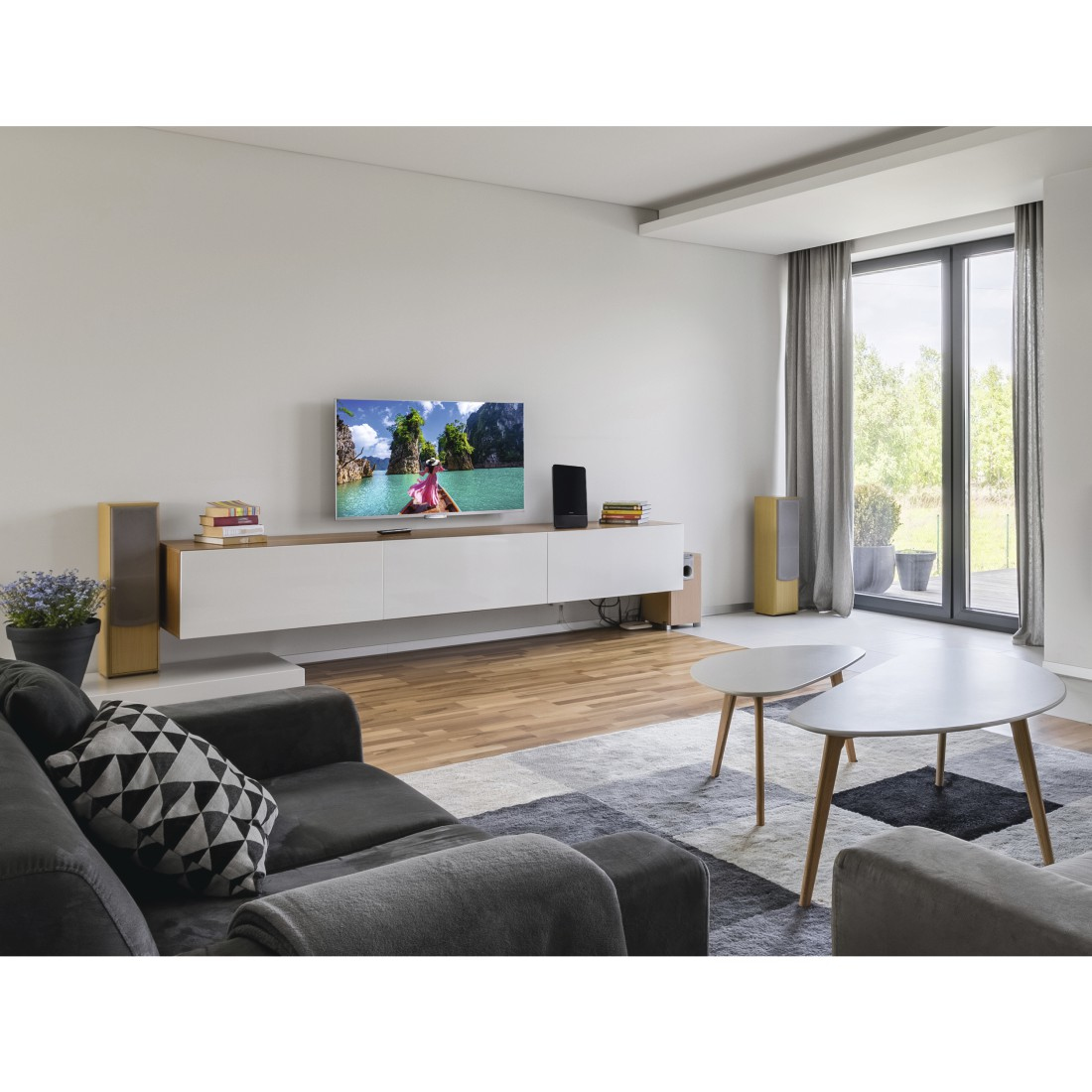 Soggiorno con divano, TV con barca sull'acqua, altoparlanti, tavolini e una grande finestra con alberi all'esterno.
