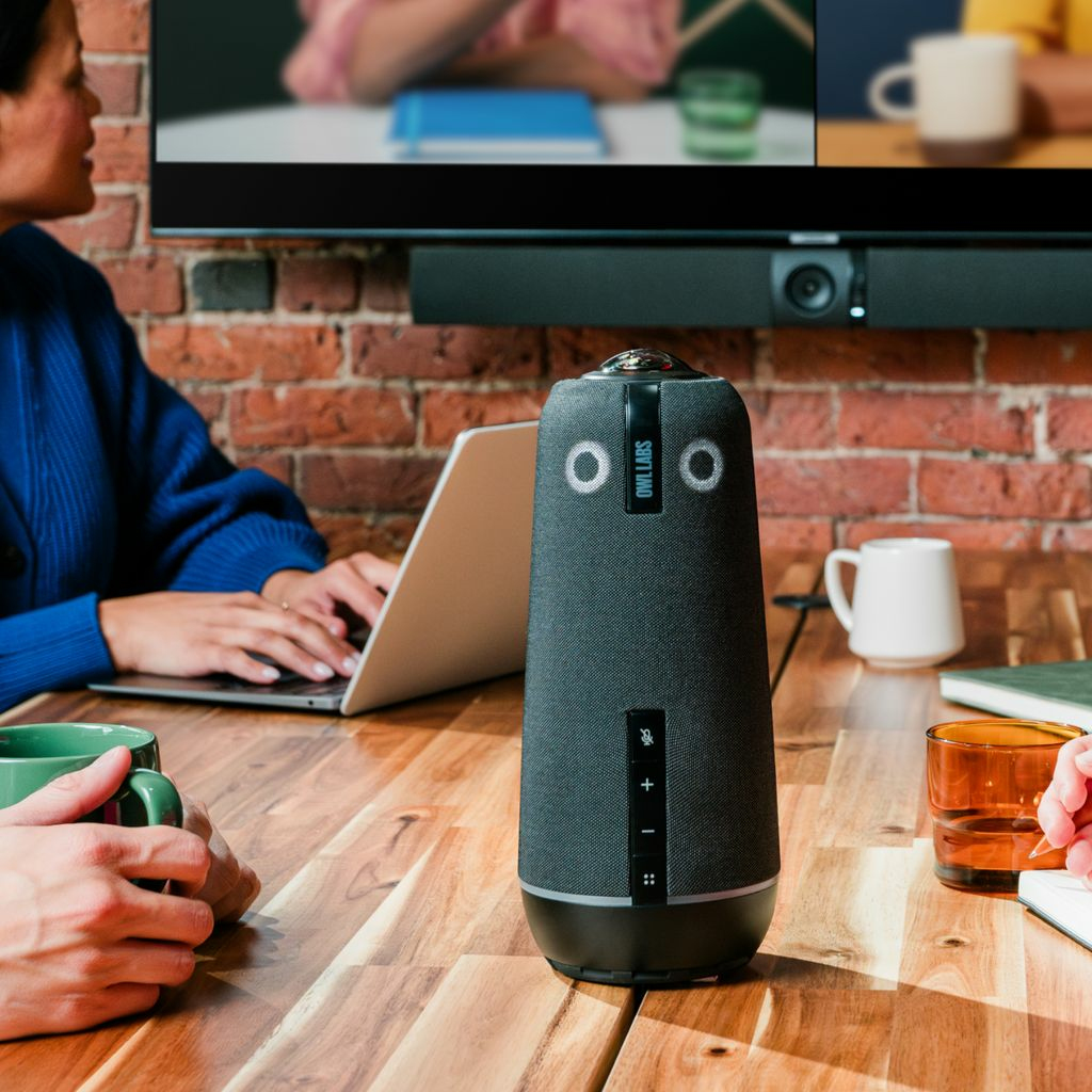 Ein Konferenzraum mit Personen, einem Laptop und einem Owl Labs Lautsprecher. Ein Fernseher ist an einer Backsteinmauer befestigt.