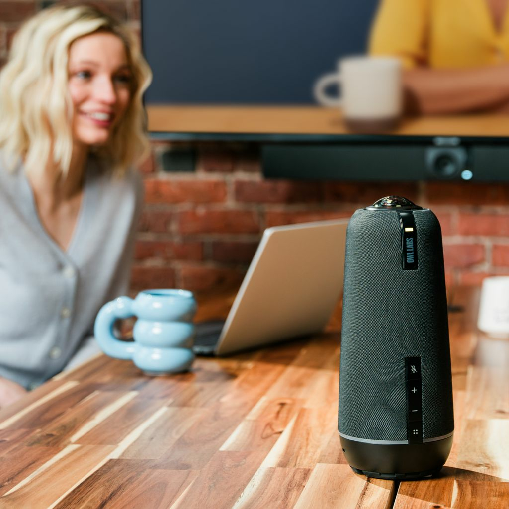 Eine Frau lächelt in der Nähe eines Lautsprechers. Auf einem Holztisch befinden sich ein Laptop, eine blaue Tasse und eine Ziegelmauer. Im Hintergrund ist eine Person zu sehen.