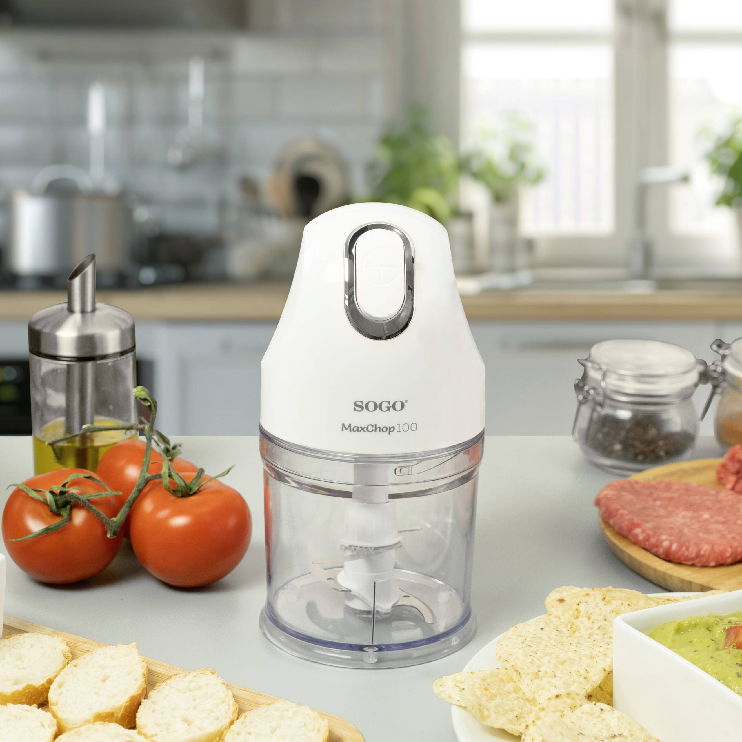 Un robot da cucina bianco su un tavolo con pomodori, pane, guacamole e patatine. Lo sfondo è una cucina.