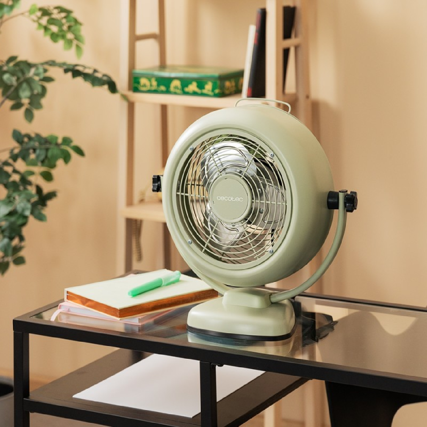 Un ventilador verde sobre una mesa de cristal con cuadernos y un bolígrafo. Un estante de madera al fondo con libros y una caja verde.