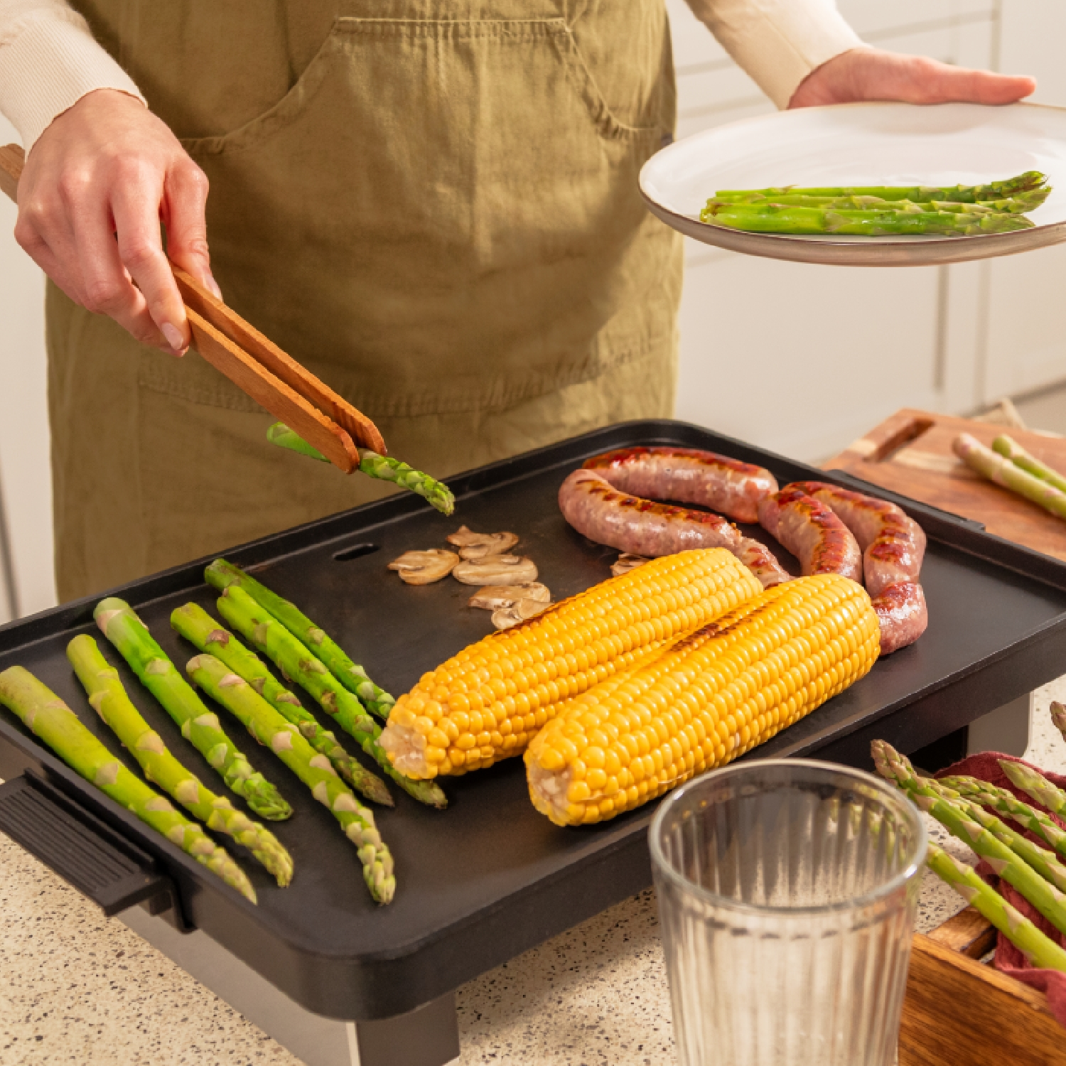 Una persona griglia asparagi, mais, salsicce e funghi. Una mano tiene un piatto di asparagi, un'altra usa le pinze.
