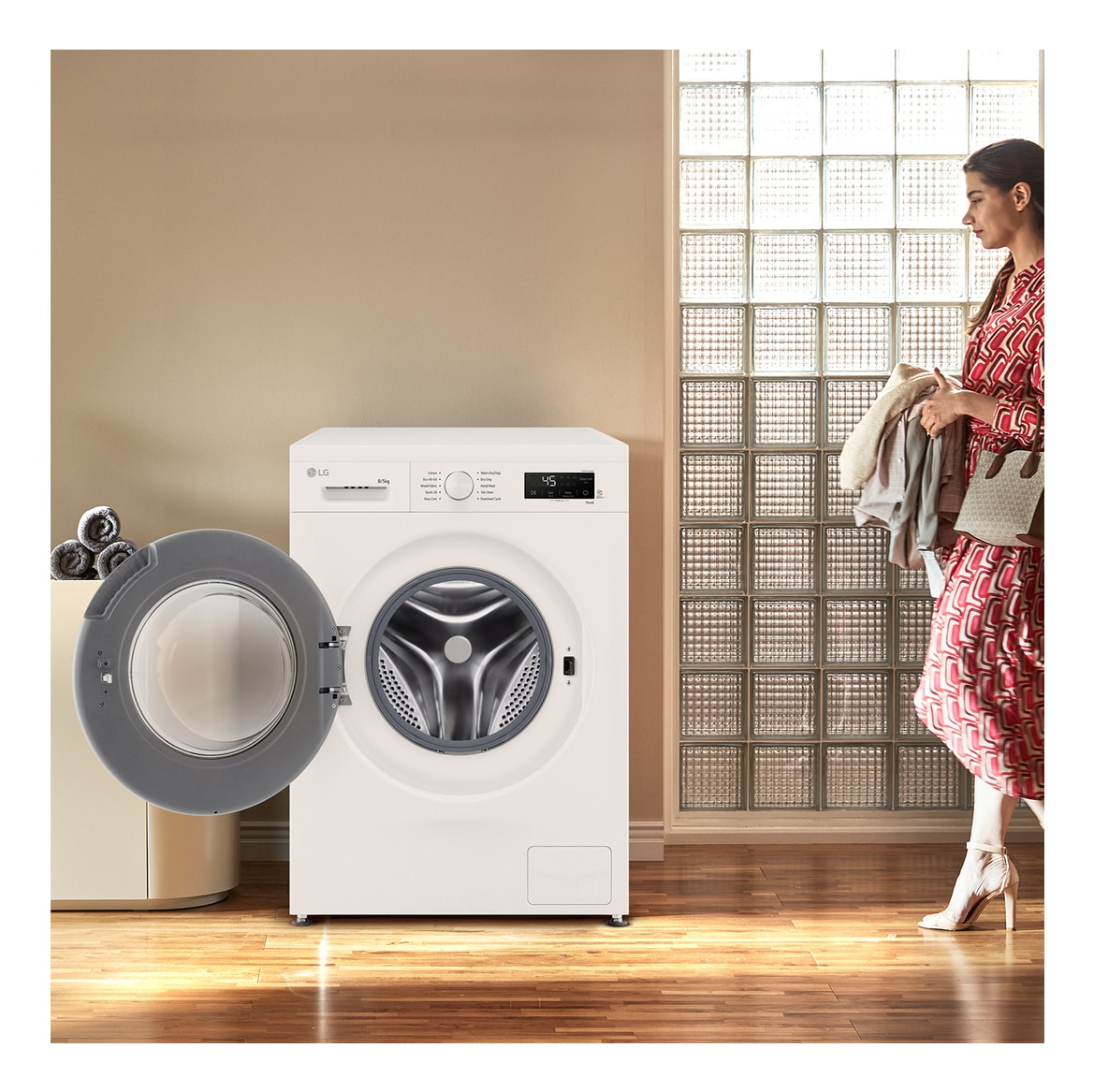 Kobieta w czerwonej sukience spaceruje w pobliżu białej pralki LG z otwartymi drzwiami. Tło ma szklane bloki.