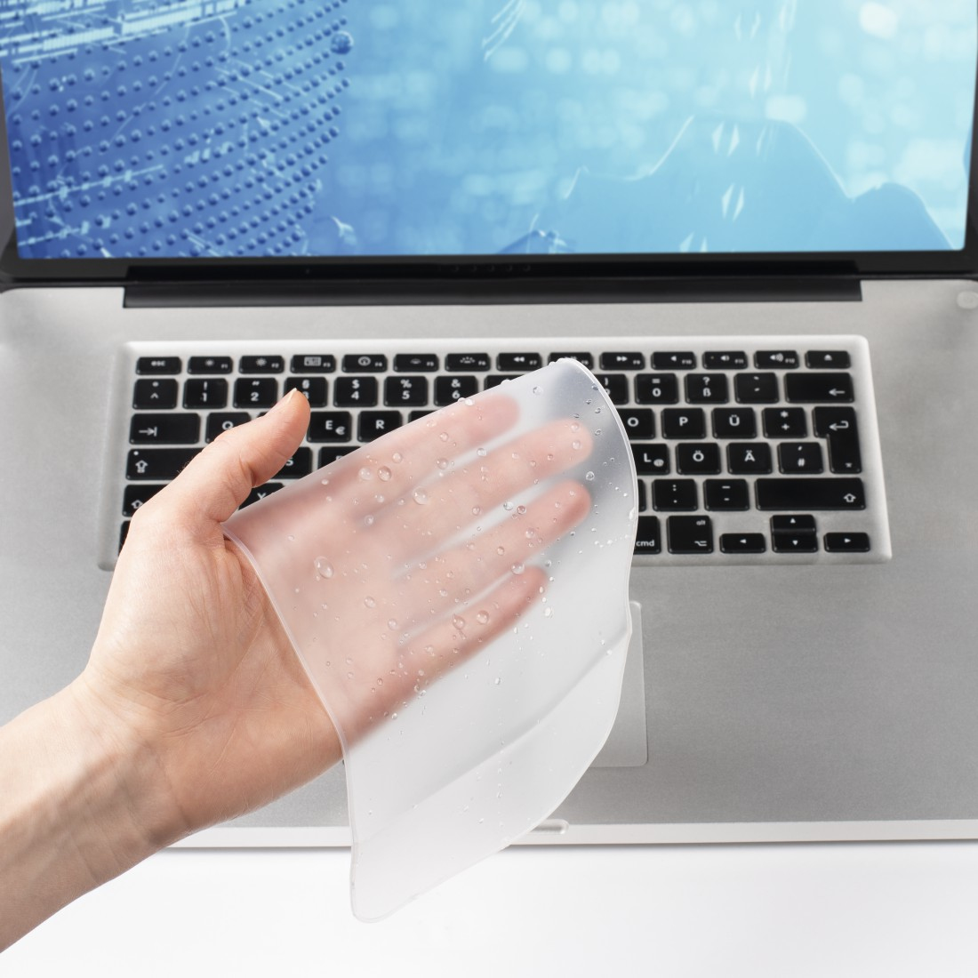 Eine Hand hält eine transparente, nasse Tastaturabdeckung über einem Laptop. Die Tastatur ist darunter sichtbar, im Hintergrund ein Bildschirm.