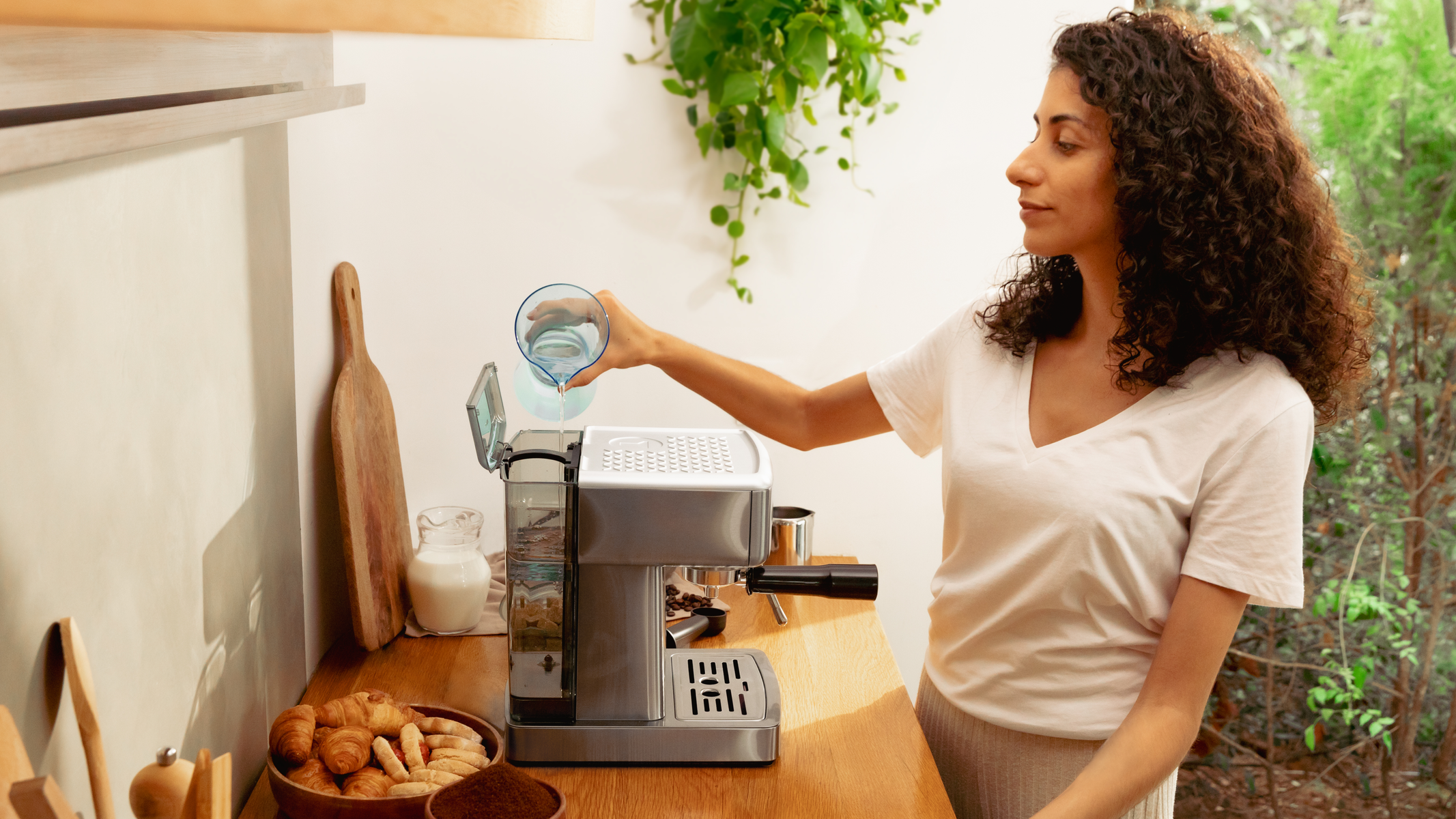 Kobieta wlewa wodę do ekspresu do kawy. W pobliżu rogaliki, mleko i ziarna kawy. Zielone rośliny w tle.