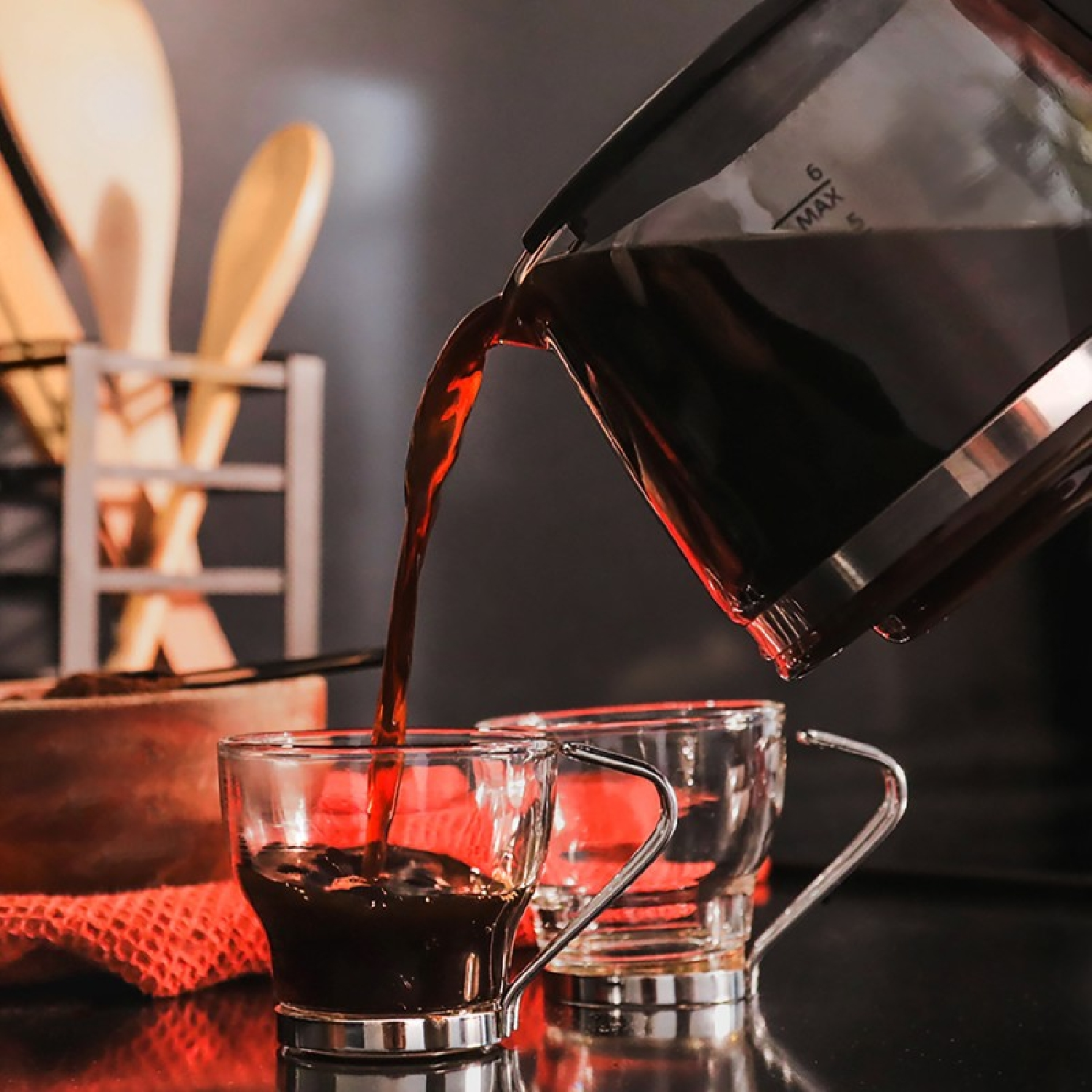 Kawa jest wlewana do dwóch przezroczystych szklanek. Drewniana łyżka i inne przybory kuchenne znajdują się w tle.