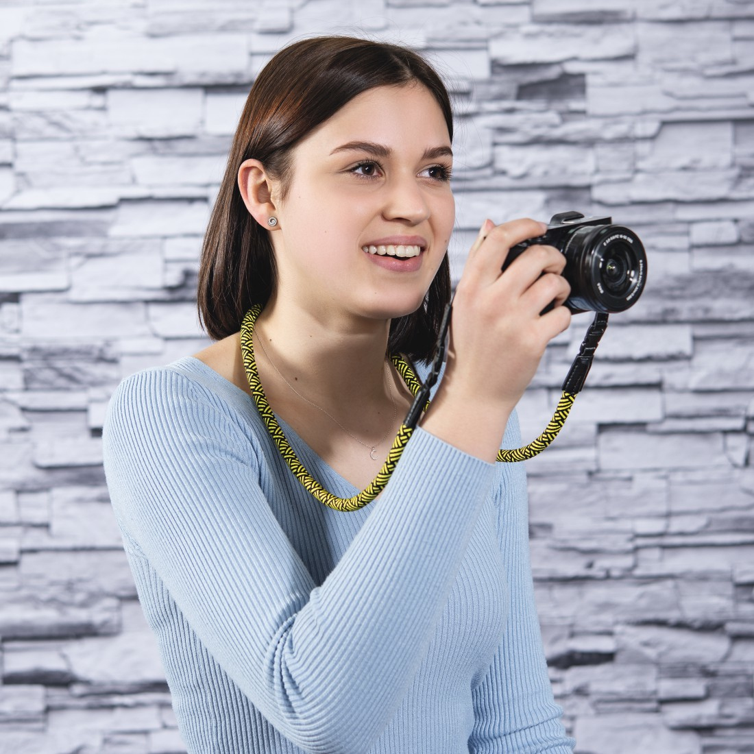 Uśmiechnięta młoda kobieta trzyma aparat z żółto-czarnym paskiem. Ma na sobie niebieski sweter na tle kamiennej ściany.