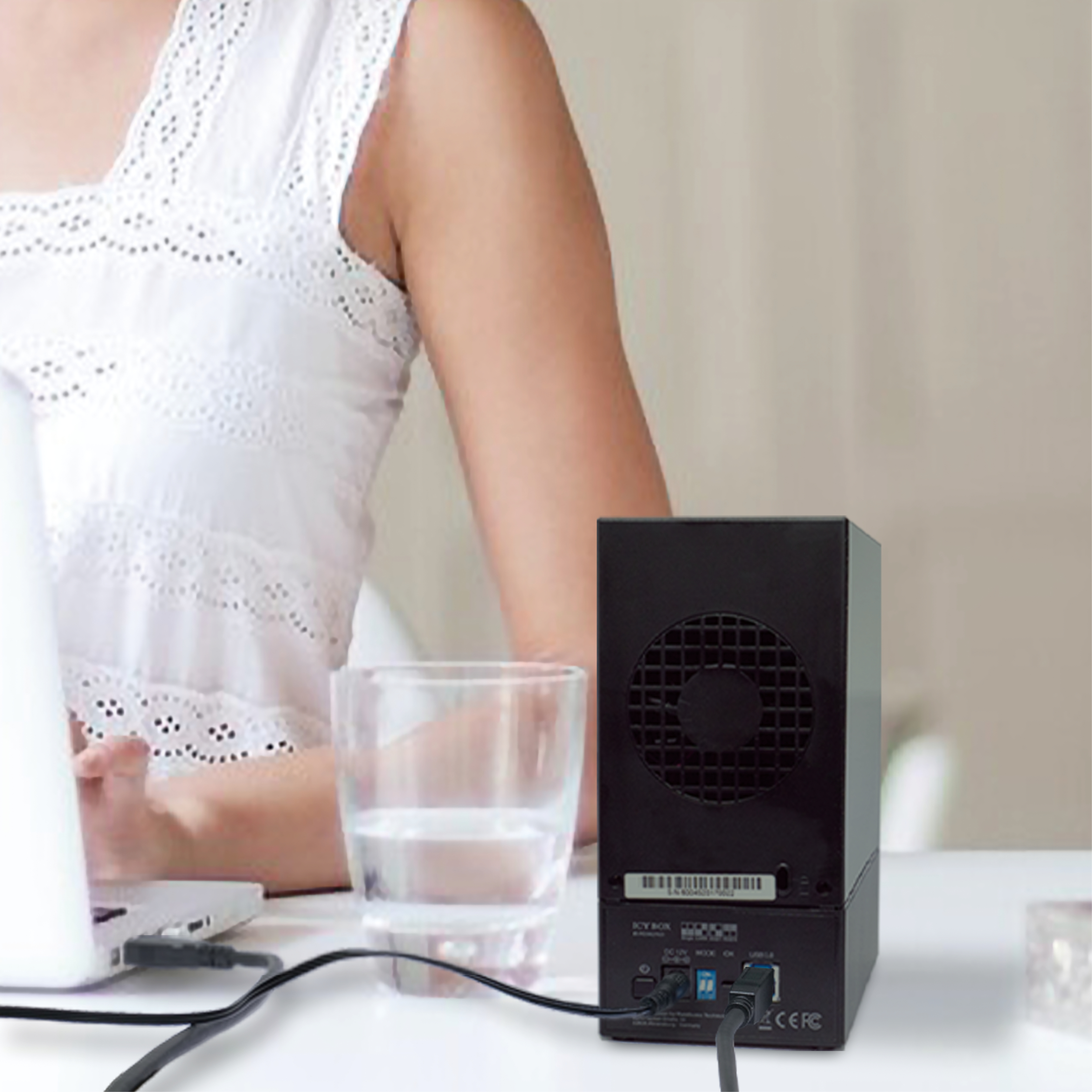 Eine Frau sitzt mit einem Laptop und einem schwarzen Gerät. Ein Glas Wasser befindet sich vor dem Gerät. Kabel sind angeschlossen.
