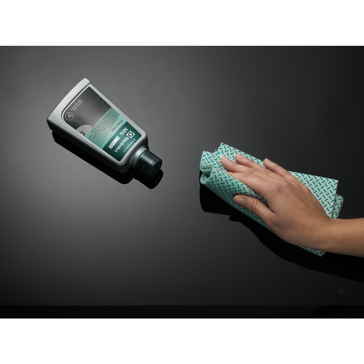 Ręka wyciera czarną powierzchnię zieloną wzorzystą szmatką. Z lewej strony stoi butelka środka czyszczącego.