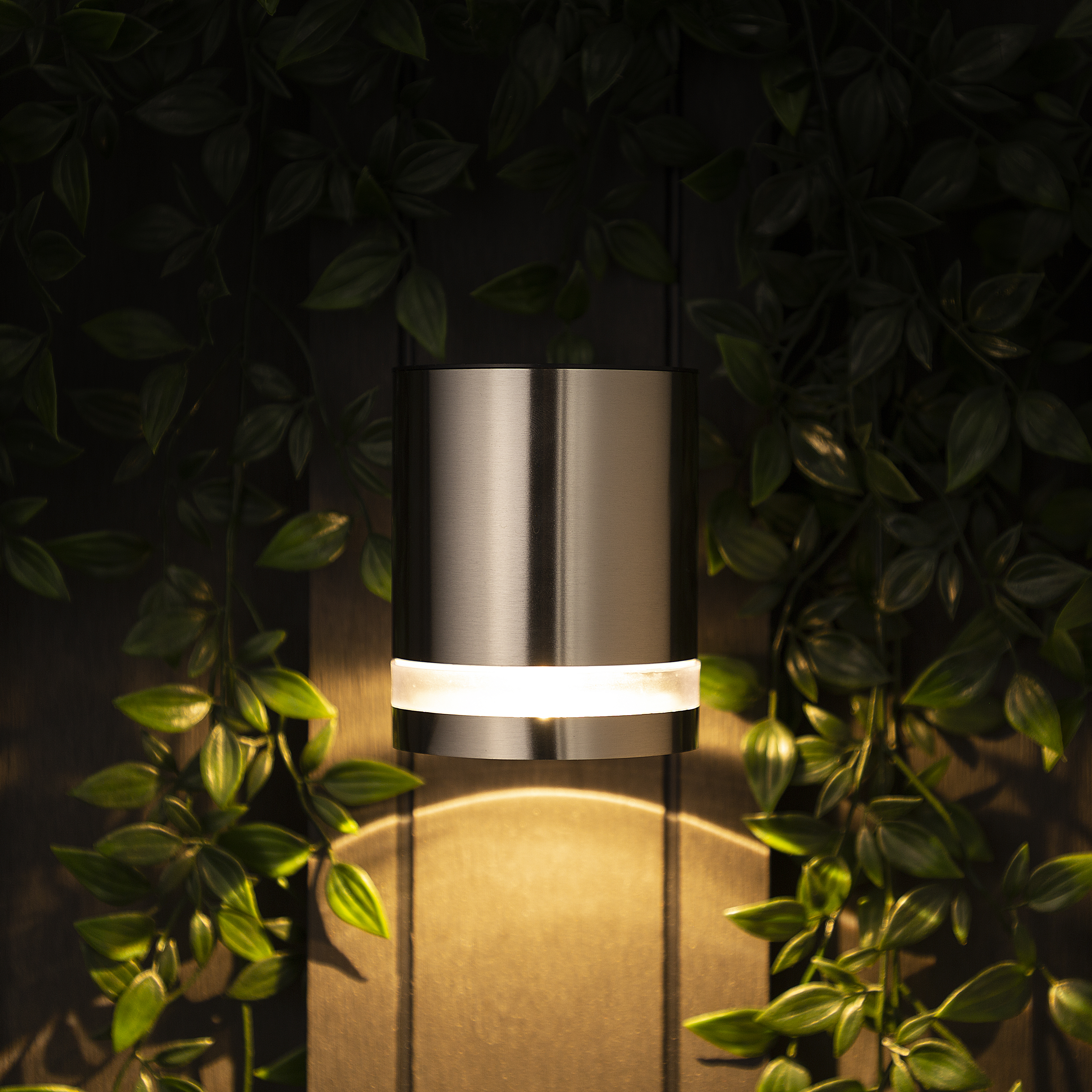 Cylindryczna, metalowa lampa ścienna emituje światło. Otaczają ją zielone liście rośliny, na tle drewna.