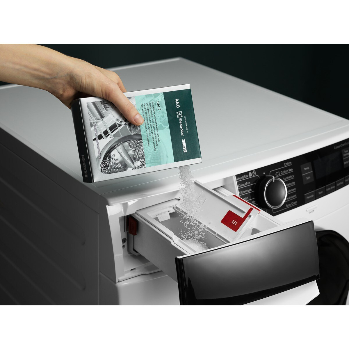 Ręka wsypuje sól z pudełka do szuflady na detergent w pralce. Maszyna jest biała z czarnymi akcentami.