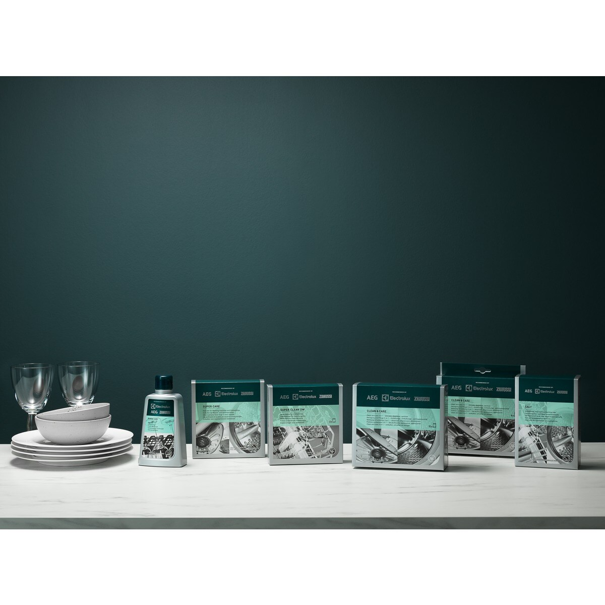 Produkty do zmywarek i zastawa stołowa na białej powierzchni. Pudełka z tekstem i obrazami pośrodku, talerze i szklanki po lewej.