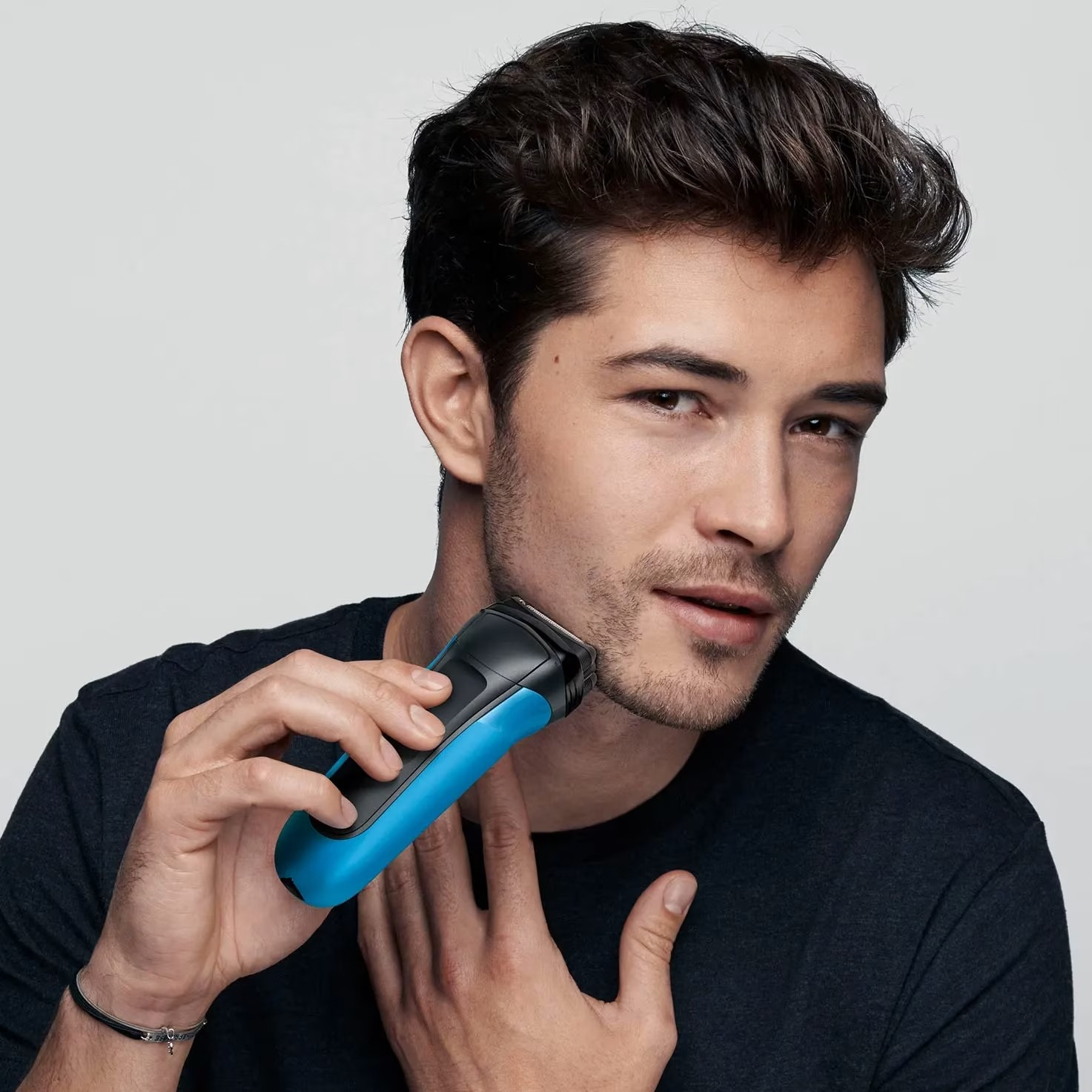 Un homme se rase le visage avec un rasoir électrique. Le rasoir est bleu et noir. Il porte une chemise noire, a les cheveux foncés et regarde la caméra.