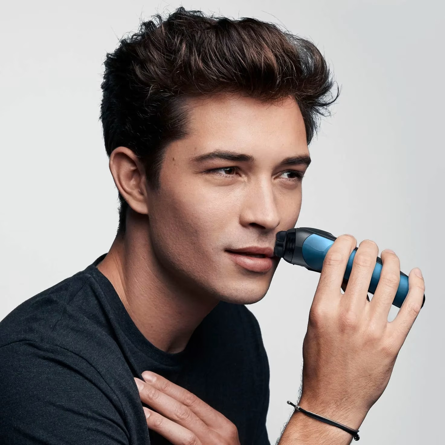 Un homme se rase le visage avec un rasoir électrique bleu et noir. Il porte un t-shirt noir et un bracelet, sur fond blanc.