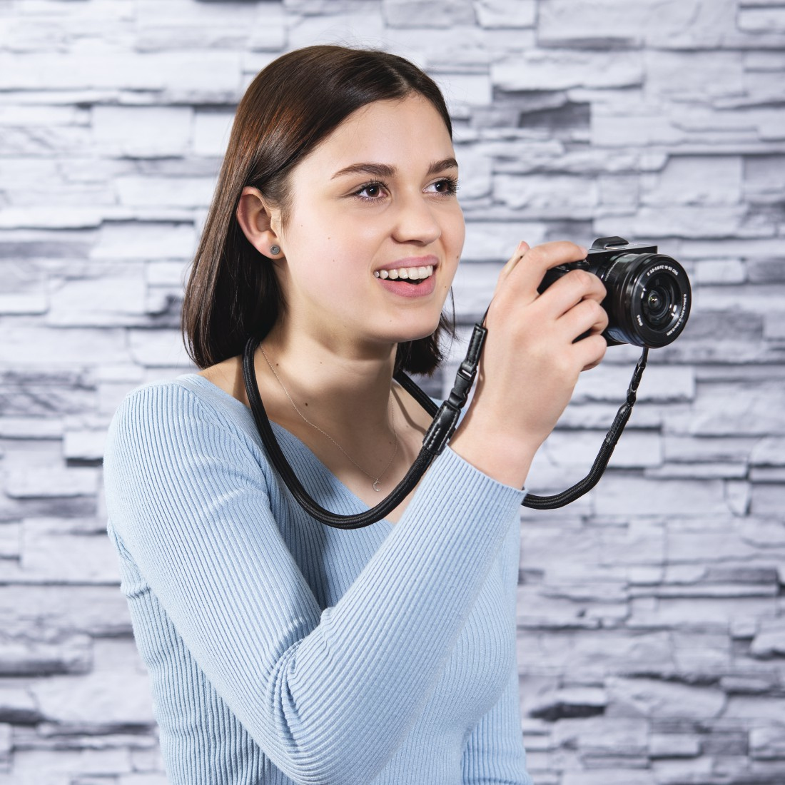 Uśmiechnięta kobieta trzyma czarny aparat z paskiem. Ma na sobie jasnoniebieski sweter. Tło to szara ceglana ściana.