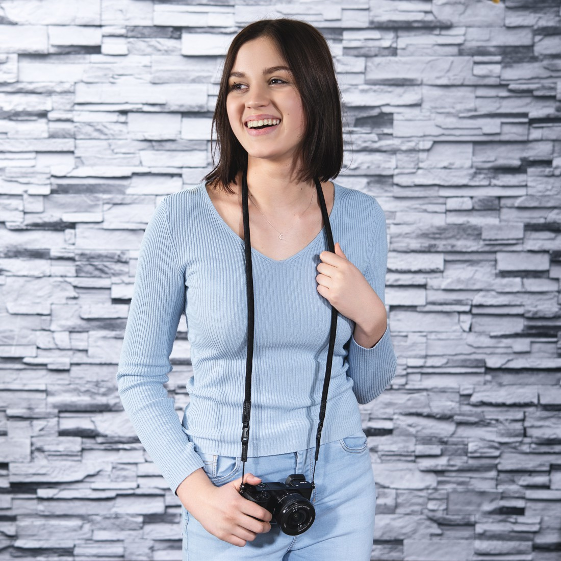 Uśmiechnięta kobieta trzymająca aparat. Ma na sobie niebieski sweter i dżinsy. Tłem jest szara kamienna ściana.