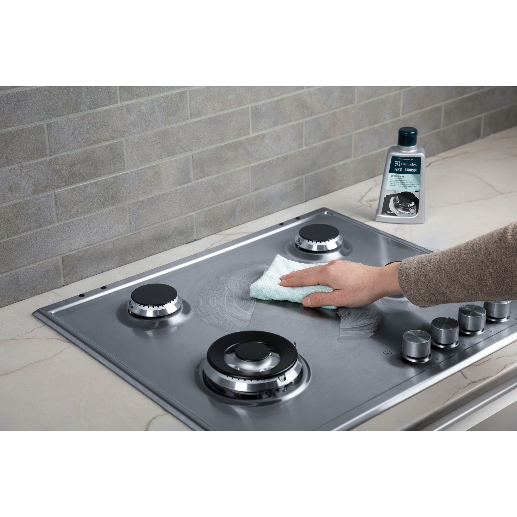 Osoba czyści kuchenkę gazową ze stali nierdzewnej zieloną szmatką. Na kuchence stoi butelka środka czyszczącego.