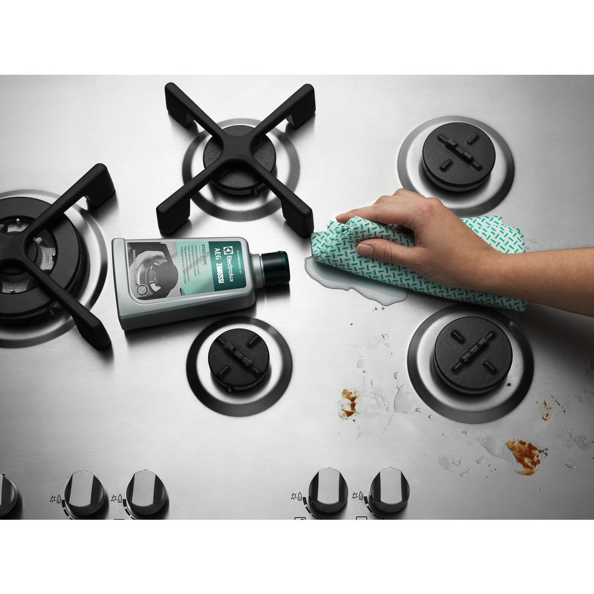 Ręka wyciera ze stali nierdzewnej kuchenkę zieloną szmatką. Obecne są plamy i butelka z płynem czyszczącym.