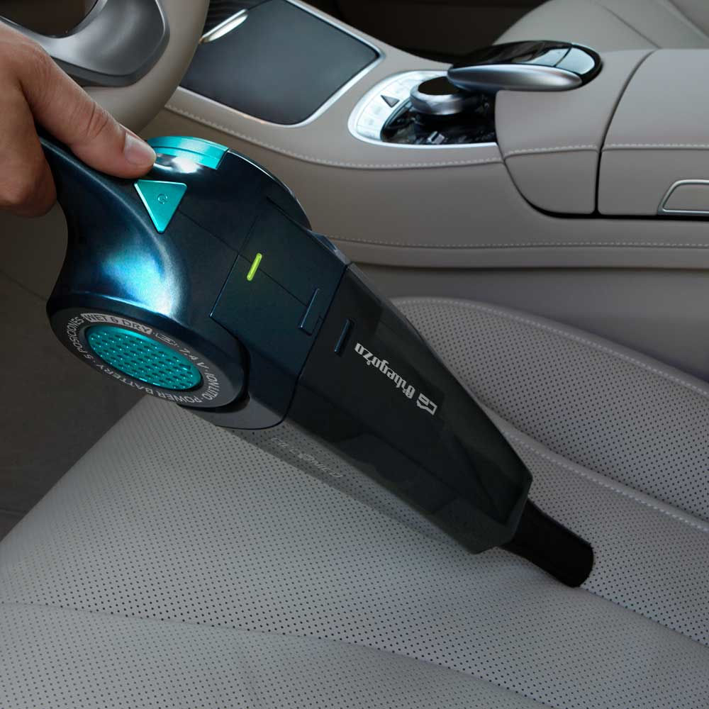 Una mano sostiene una aspiradora de coche negra y verde azulado sobre un asiento de coche de cuero blanco. Interior de un coche en el fondo.