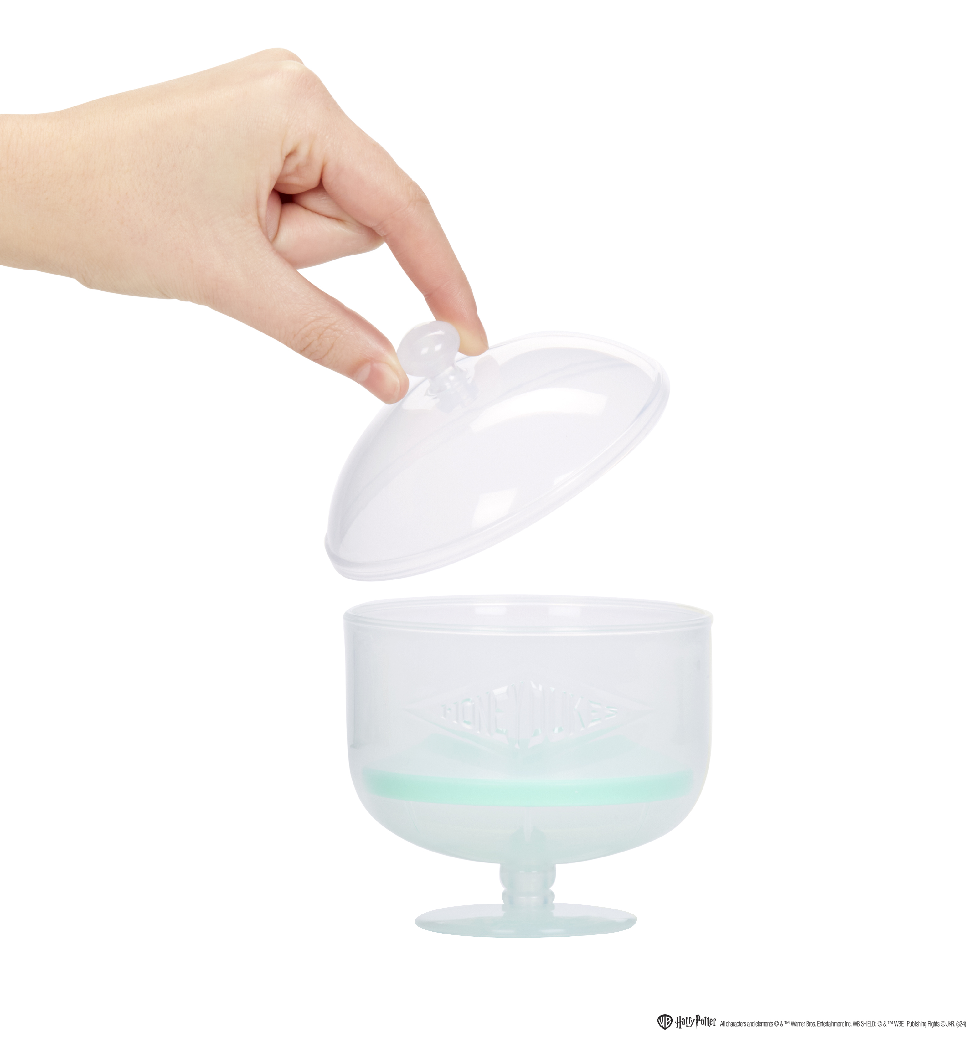 Ręka otwiera przezroczysty pojemnik na cukierki z pokrywką. Ma miętową podstawę i napis HONEYDUKES.