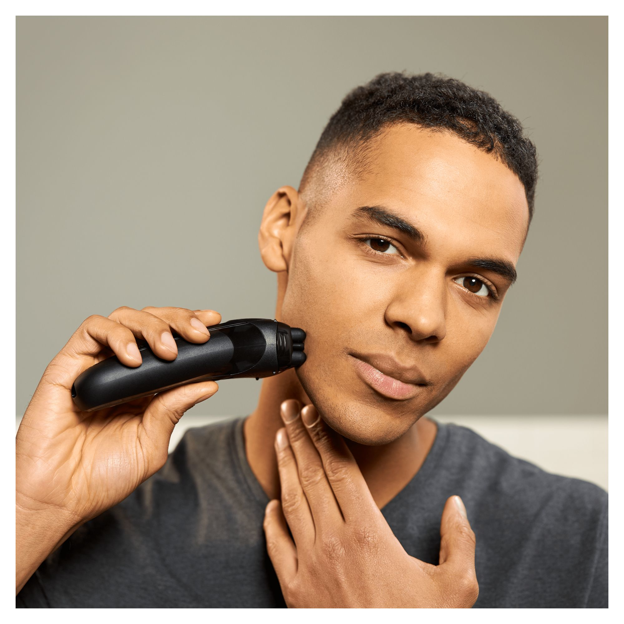 Mężczyzna goli twarz czarną golarką elektryczną. Dotyka szyi drugą dłonią. Neutralne tło.