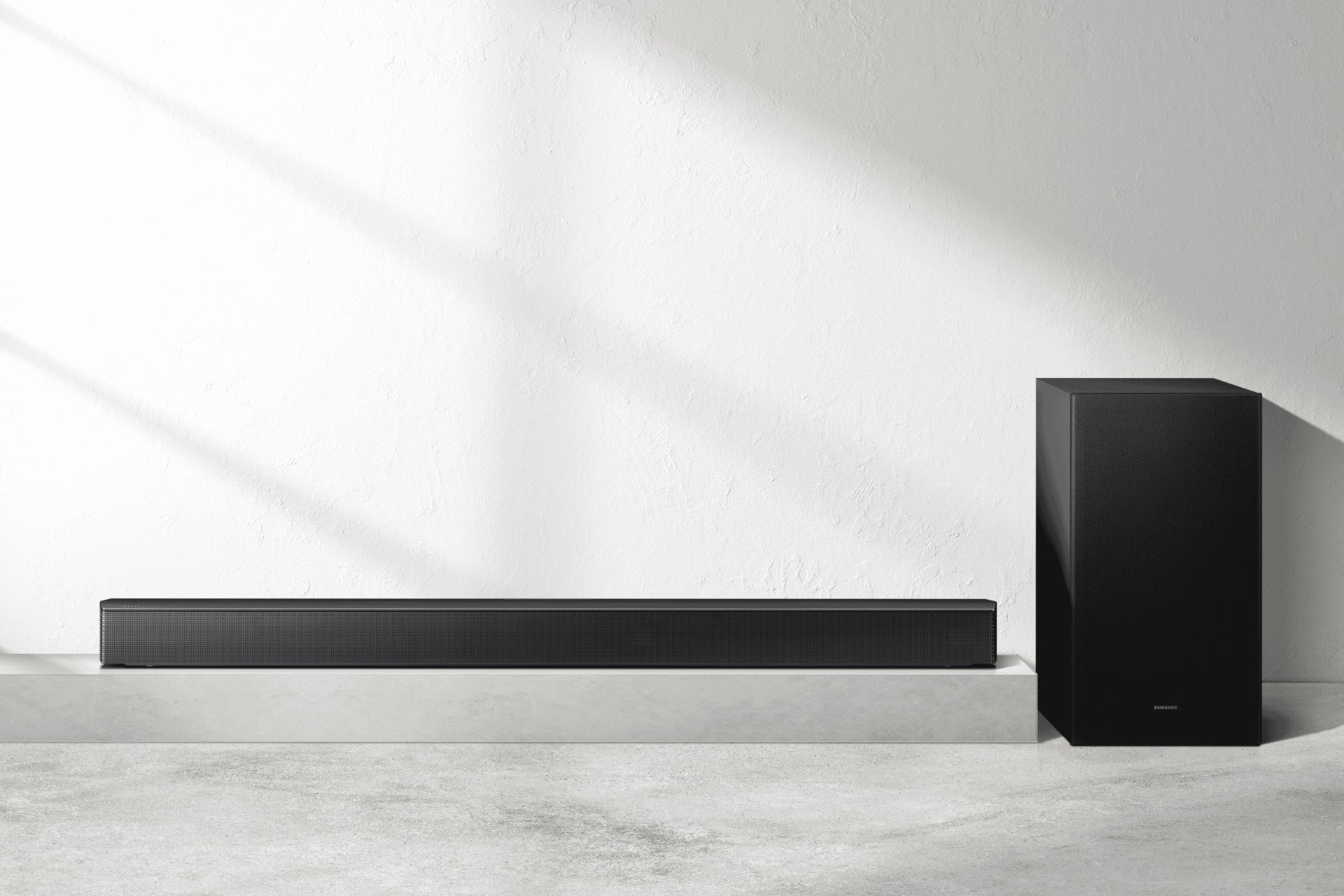 Eine schwarze Soundbar und ein Subwoofer stehen auf einer grauen Oberfläche vor einer weißen, strukturierten Wand. Schatten werden geworfen.