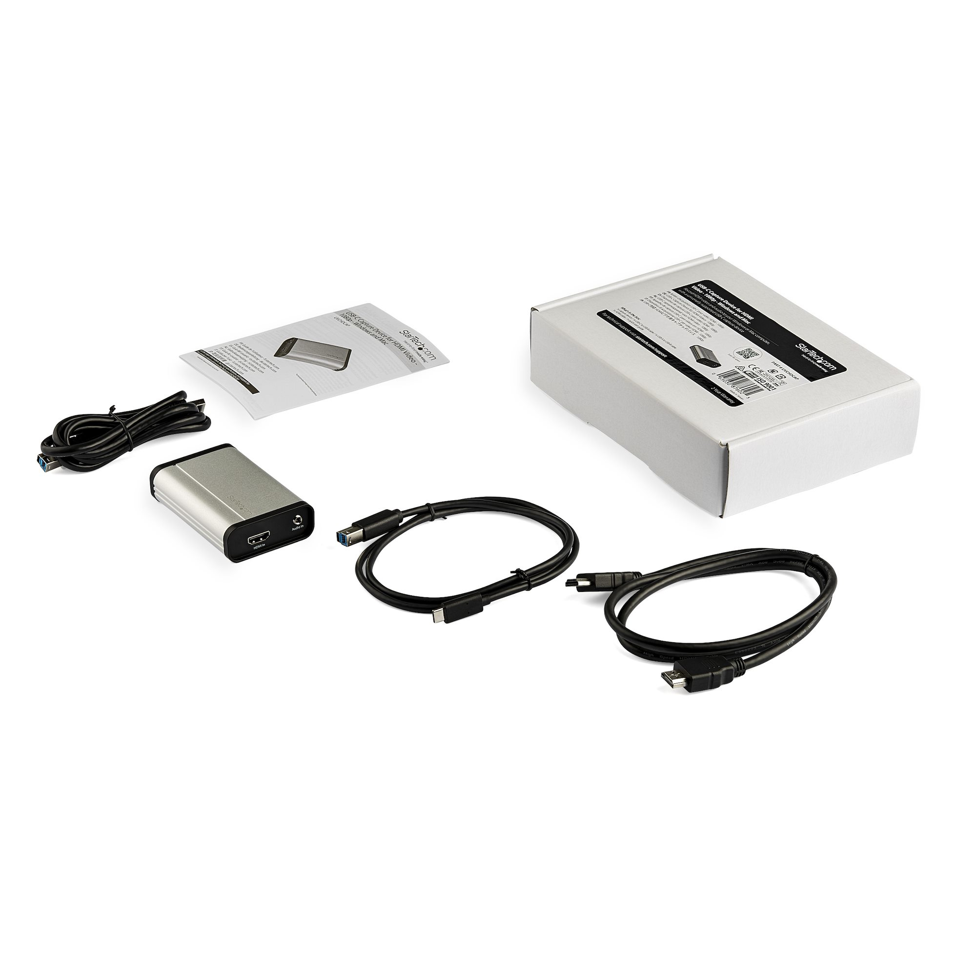 Dispositivo de captura plateado con cables, caja y documentos sobre una superficie blanca. La caja y el dispositivo tienen el logo StarTech.com.